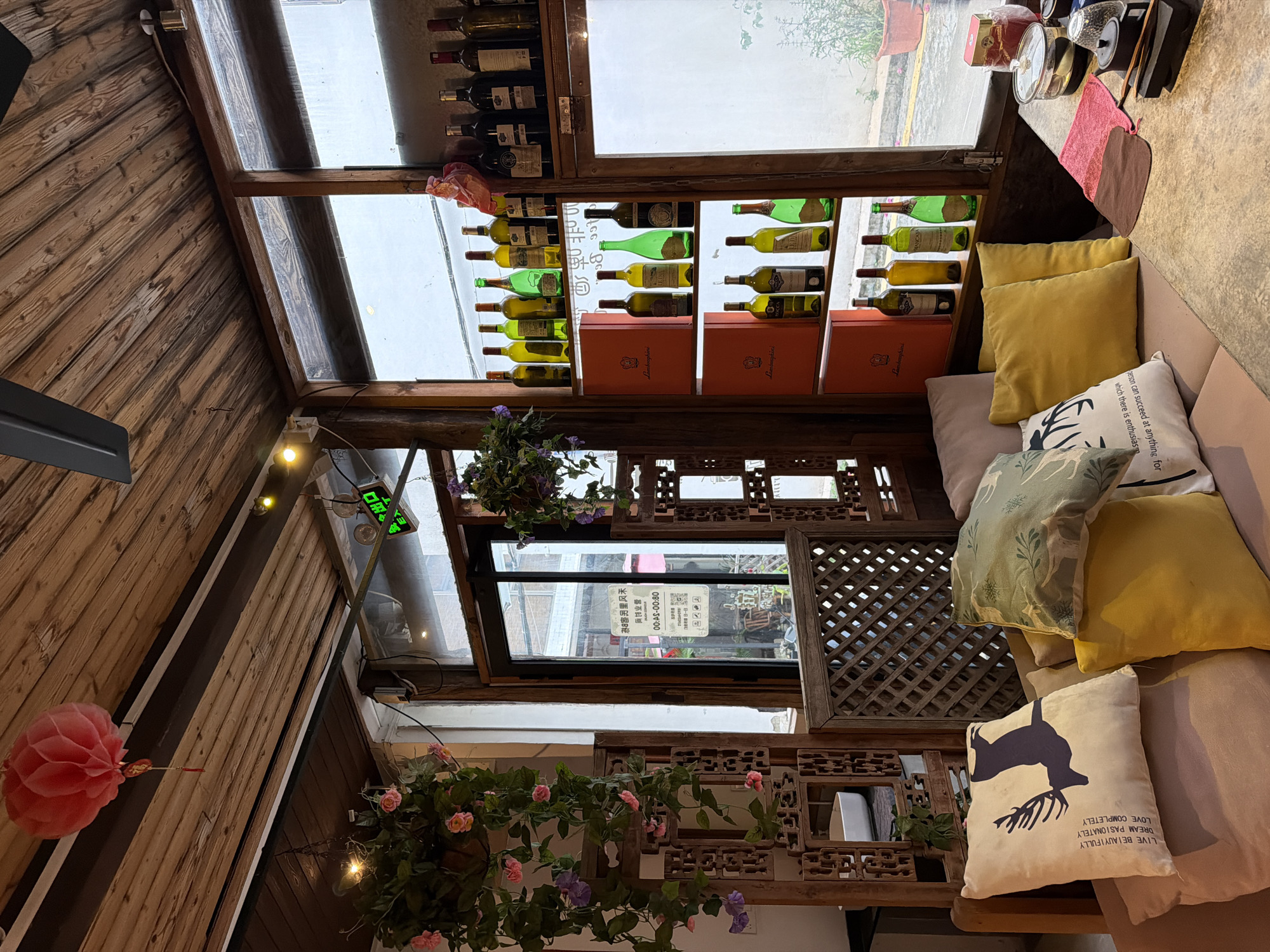 The image depicts the interior of a cozy establishment, possibly a cafe or wine bar, located in Longgang District, China. The room features wooden paneled walls and ceiling, with a decorative red lantern hanging from the ceiling. Various potted plants with green leaves and pink flowers are arranged around the space, adding a touch of nature.
In the foreground, a sofa is adorned with several cushions. One cushion on the left features a dark silhouette of a deer with antlers and text in English. To its right, another cushion has a pattern of leaves and vines. Two yellow cushions are placed in front of these.
The background reveals a large window showcasing shelves filled with wine bottles. The bottles are of various shapes and colors, suggesting a diverse collection. Some are green, some clear, and some dark. Orange boxes with a red logo are stacked on the shelves below the bottles. Adjacent to the shelves is a glass door with a sign that appears to be a business opening hours notice in Chinese.
On a table in the right side of the image, there are several items, including what looks like teacups, a glass, and a container with a lid, possibly for tea or snacks. A pink cloth and a brown cloth are also placed on the table. The overall ambiance is warm and inviting, with a rustic yet modern aesthetic. There are no people visible in the image.