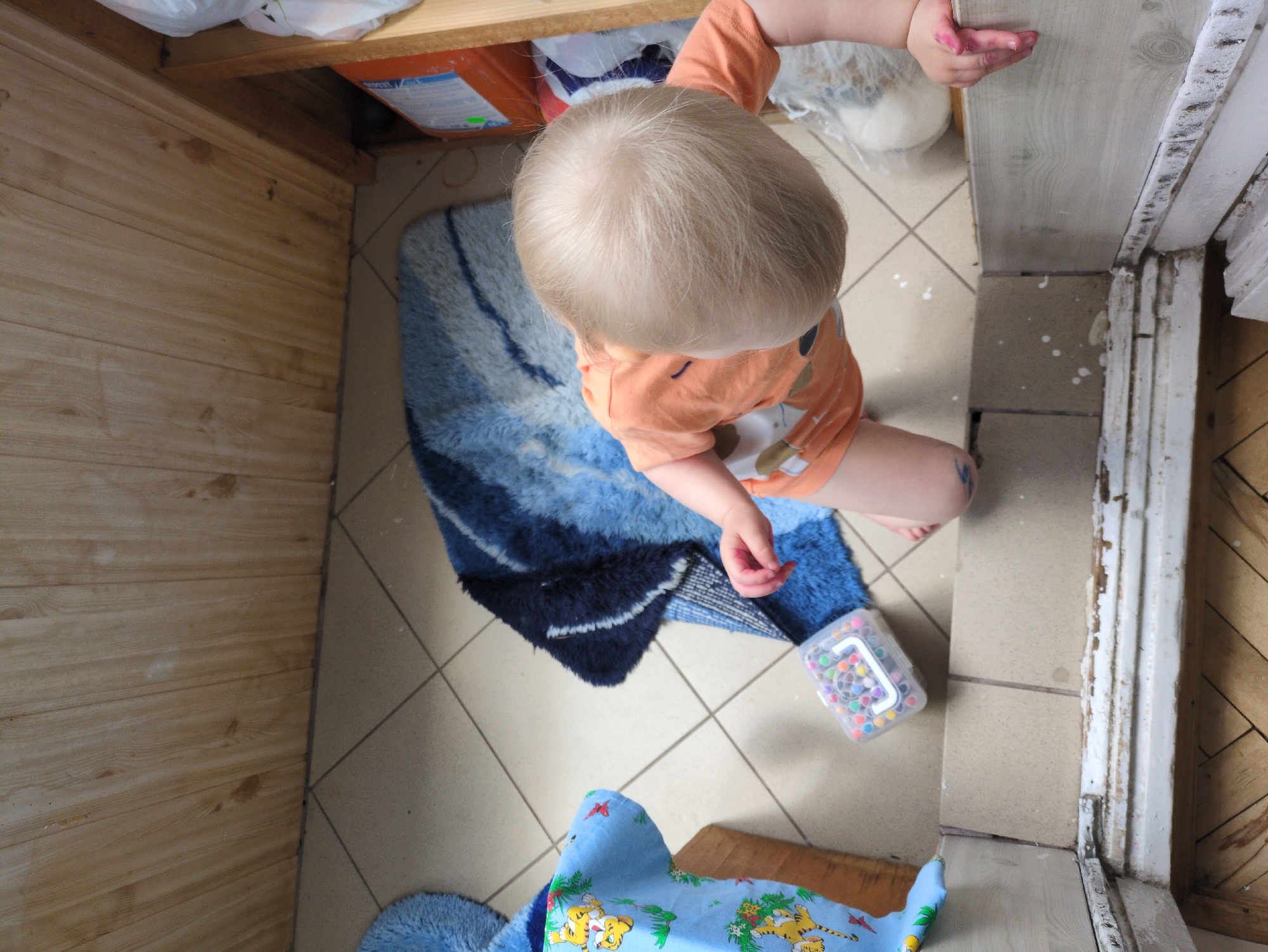 This is a high-angle, close-up, indoor photograph taken in Dubliany, Ukraine. The scene depicts a toddler, appearing to be between 1-2 years old, with blonde hair, wearing an orange t-shirt with a cartoon animal print. The child is kneeling on a tiled floor and reaching towards a white trim. The toddler's left hand has pink paint residue on it. In front of the child is a blue rug with a striped pattern. To the left of the rug is a blue fabric object with a yellow tiger print, resting on a wooden surface. A clear plastic container filled with colorful beads is positioned near the edge of the blue rug. The environment appears to be a room with wooden paneled walls and beige floor tiles. To the right of the scene, a white object, possibly a large stuffed toy, is visible partially wrapped in plastic. The overall impression is of a child engaged in a creative activity, possibly painting, in a domestic setting.