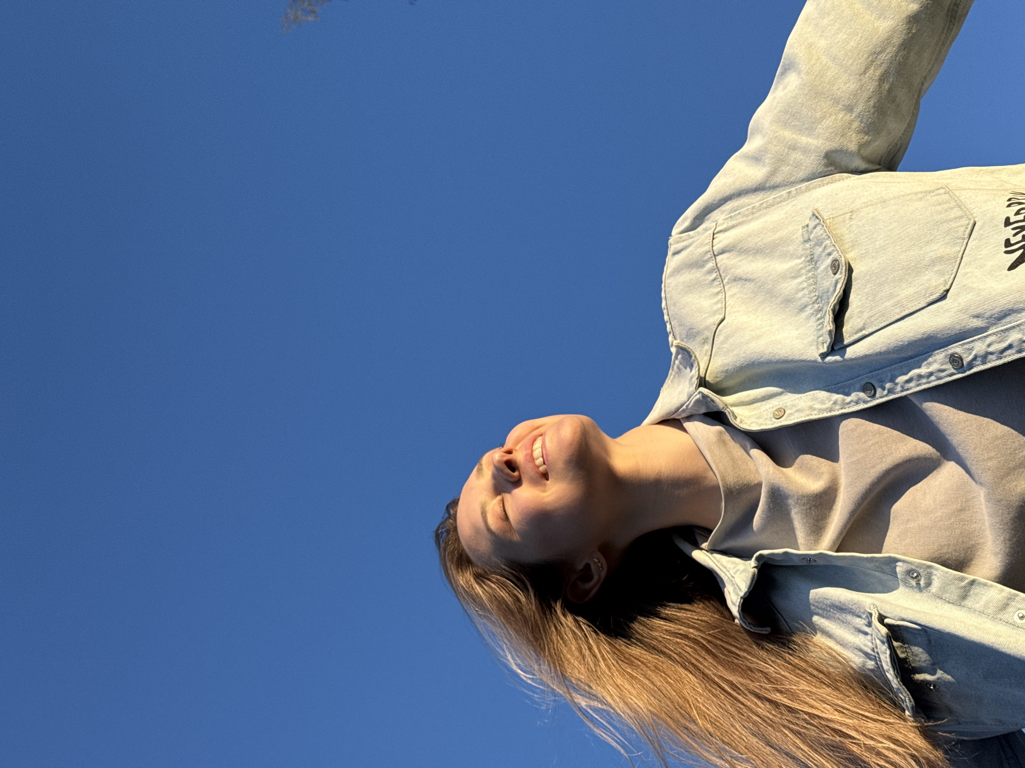 The image shows a woman with long blonde hair, wearing a light-colored denim jacket over a neutral-colored top, tilted back with her eyes closed and a smile on her face. She appears to be looking up at the sky. The background is a clear blue sky. The location, Chaikovsky, Russia, is not discernible from the image's content.