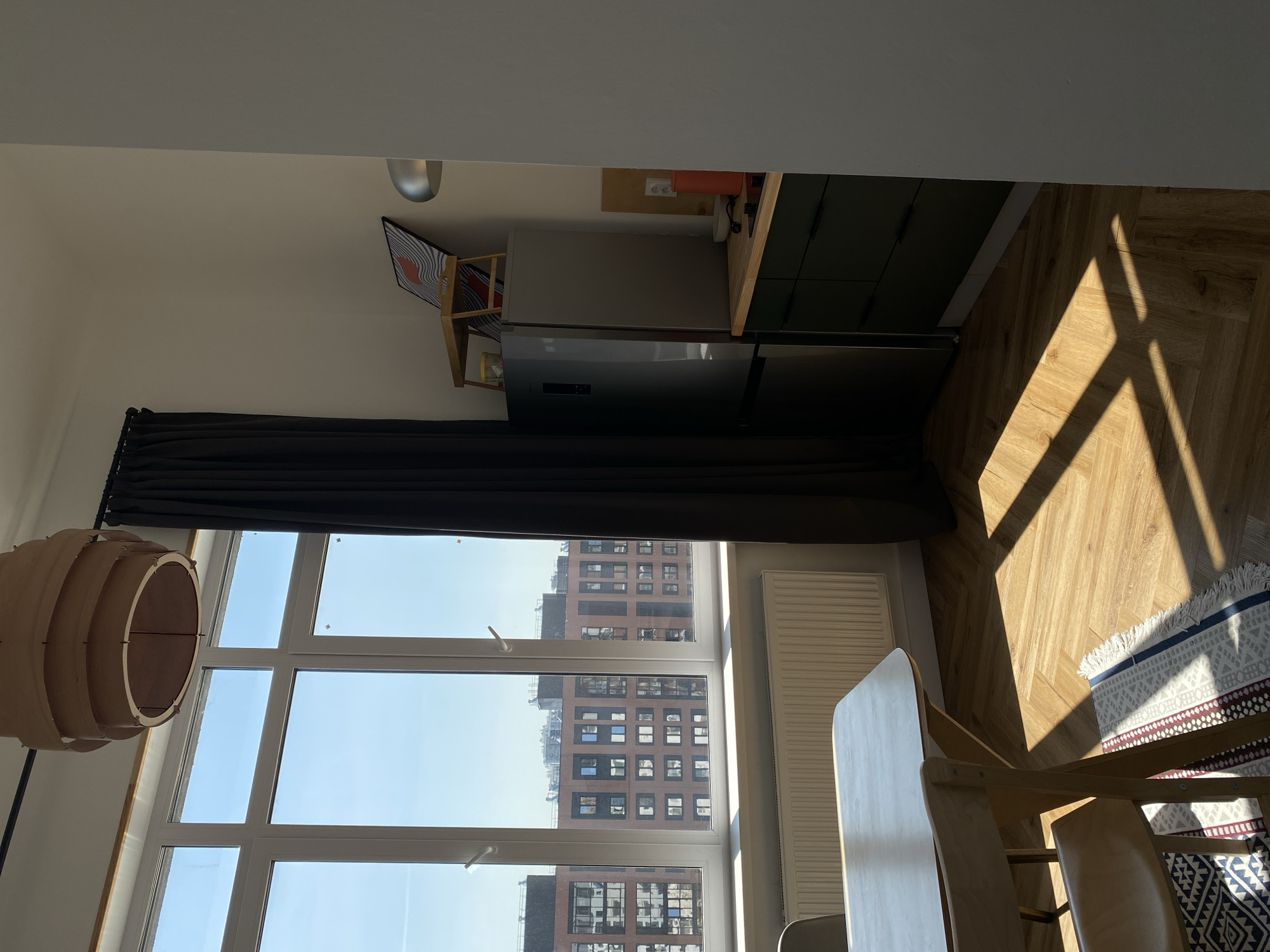 The image shows an interior view of a room in Izhevsk, Russia. The room is well-lit with sunlight streaming through a large window, which is partially covered by a dark curtain. Outside the window, tall buildings are visible, indicative of an urban environment. A wooden dining table and chairs are positioned in the room, with a rug partially visible beneath them. A unique, layered wooden lamp hangs near the window. Adjacent to the window, there's a kitchen area with a refrigerator and cabinets. The flooring is herringbone wood. The scene appears to be a residential space, possibly an apartment.