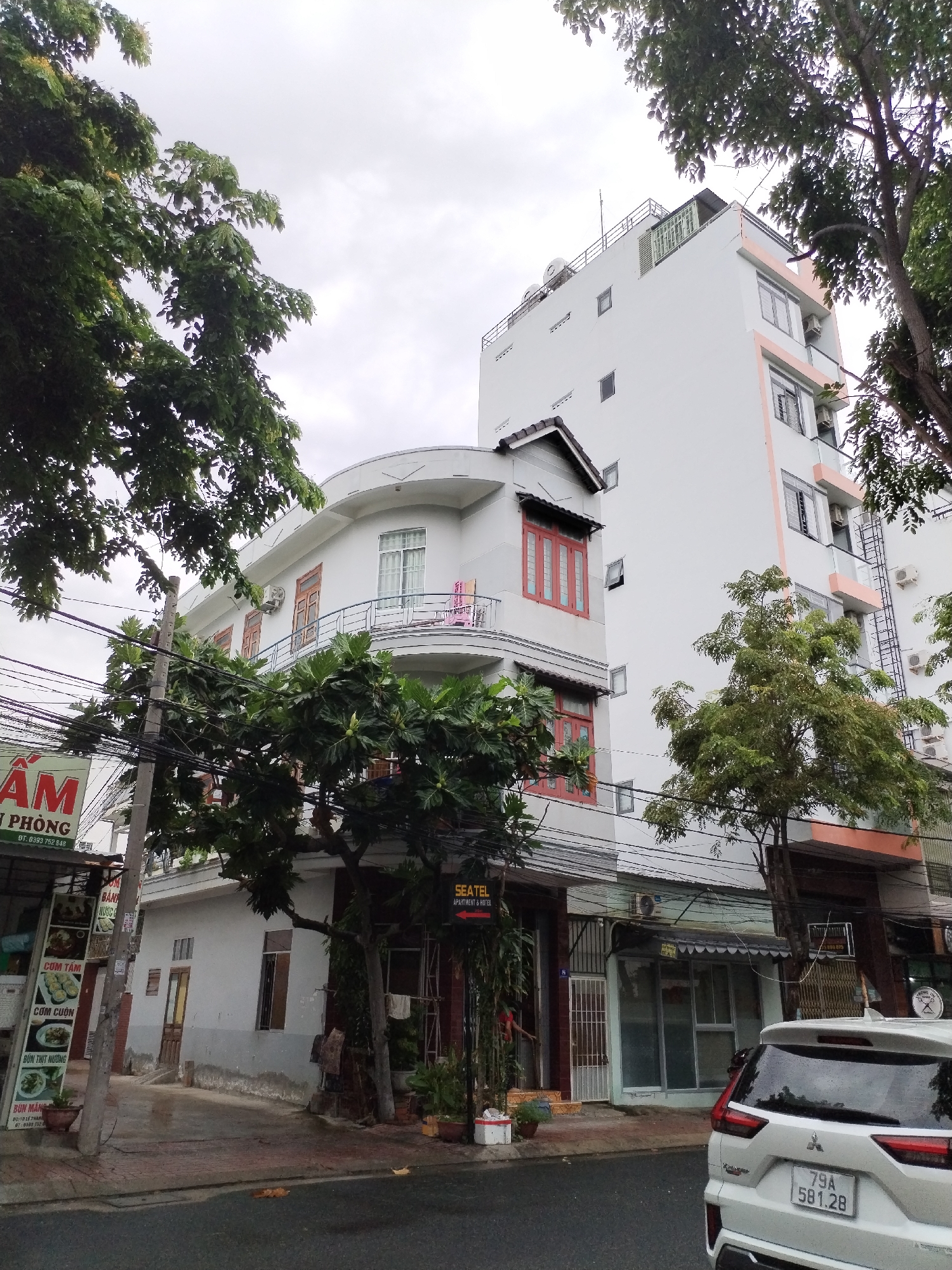 This photograph captures a daytime street scene in Nha Trang Ward, Vietnam, under an overcast sky. The focal point is a multi-story building, likely a hotel or apartment complex, featuring white facades with some sections accented by peach-colored trim and red-framed windows. The building exhibits varied architectural styles, including a section with curved balconies and another taller, more rectangular part with visible air conditioning units and a rooftop structure. Below, a ground-floor commercial space is marked with a sign for "SEATEL APARTMENT & HOTEL," near which a person is partially visible.
To the left, a street vendor's sign displays menu items, with visible text including "AM," "PHÒNG," "DT: 0393 752 848," "CƠM TÂM," "CƠM CUỐN," "BÚN THỊT NƯỚNG," and "BÚN MANG." Dense green foliage from several trees partially obscures the buildings and a tangle of overhead power lines. A white Mitsubishi Xpander SUV is parked on the wet street in the foreground, displaying the Vietnamese license plate "79A 581.28." The sidewalk shows signs of recent moisture, and the overall impression is one of a typical urban environment in Vietnam.