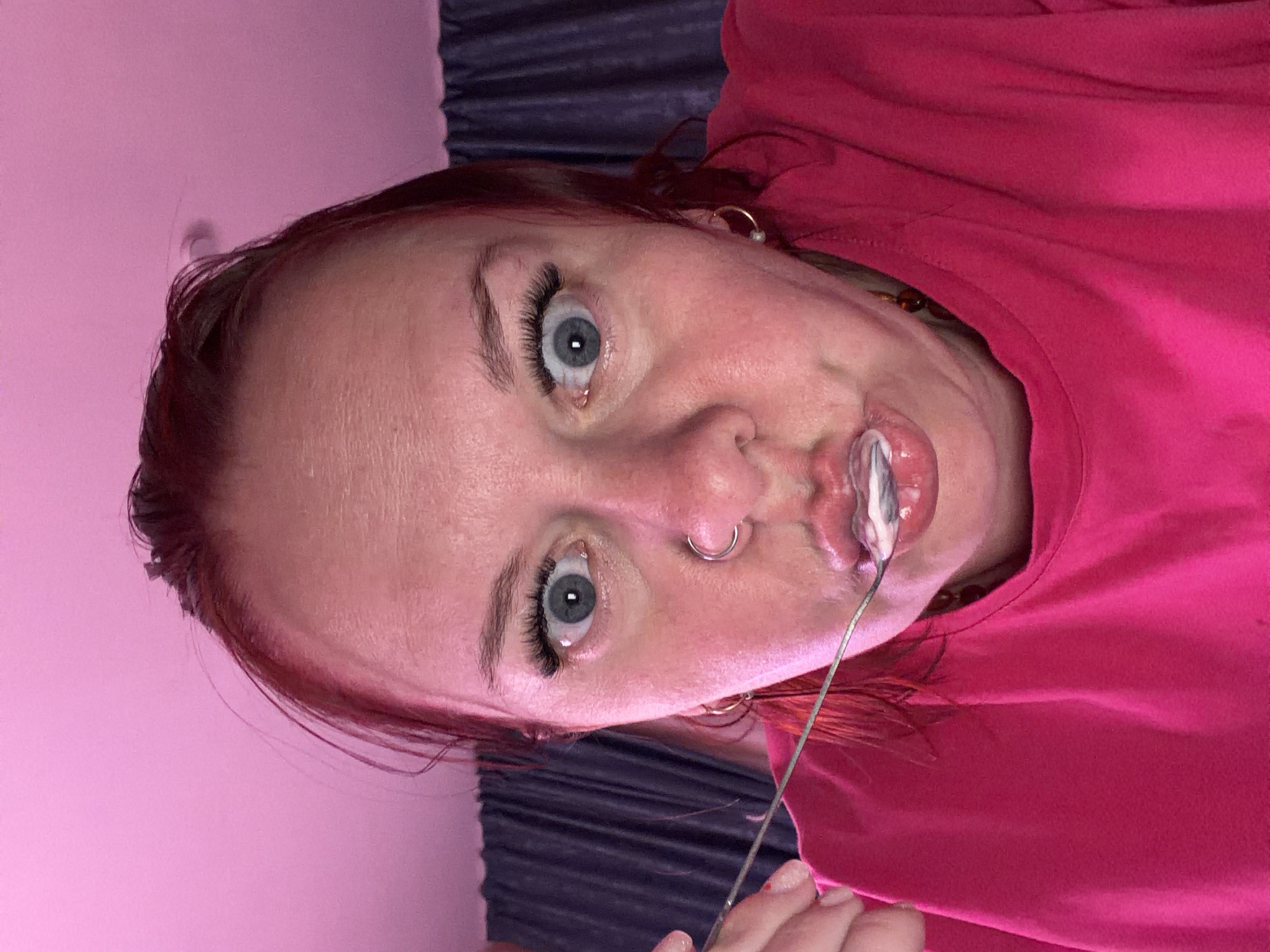 A close-up, eye-level photograph depicts a female subject indoors in Chaikovsky, Russia, consuming a white, creamy substance with a silver-colored spoon. The subject, facing forward, has fair skin with a reddish tone, reddish-brown hair, and is wearing a fuchsia-colored short-sleeved top. Her blue eyes are wide open with dilated pupils and enhanced eyelashes. A silver-colored ring is present in her septum piercing. She wears gold-colored hoop earrings: one with a white, pearl-like bead on the right, and one with a dark, amber-like bead on the left. The background features a plain pink wall or surface on the upper left, transitioning to dark blue pleated curtains on the upper and lower right. The scene is illuminated with a noticeable pink light source.