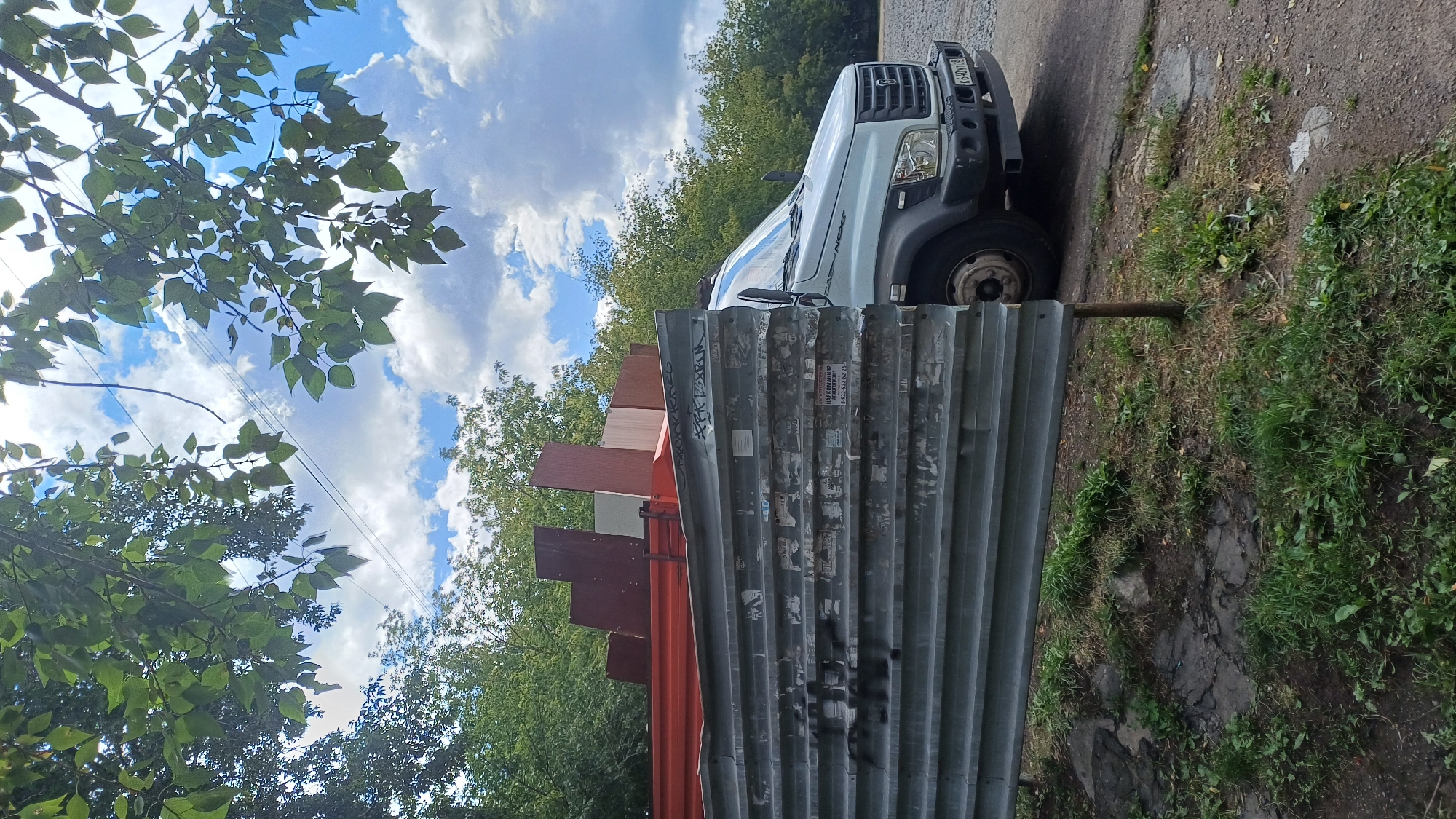 In Izhevsk, Russia, on a cloudy day, a light-colored truck, possibly a GAZ NEXT, is parked on an asphalt surface adjacent to a patch of unkempt grass. The truck is loaded with reddish-brown and light-colored rectangular panels that appear to be construction materials or furniture. In the foreground, a corrugated metal fence, partially obscured by leaves, runs horizontally across the frame. The fence is weathered and covered in various stickers and graffiti. Overhead, branches with green leaves are visible against the sky, with power lines crisscrossing between them. The general environment suggests an outdoor industrial or storage area.