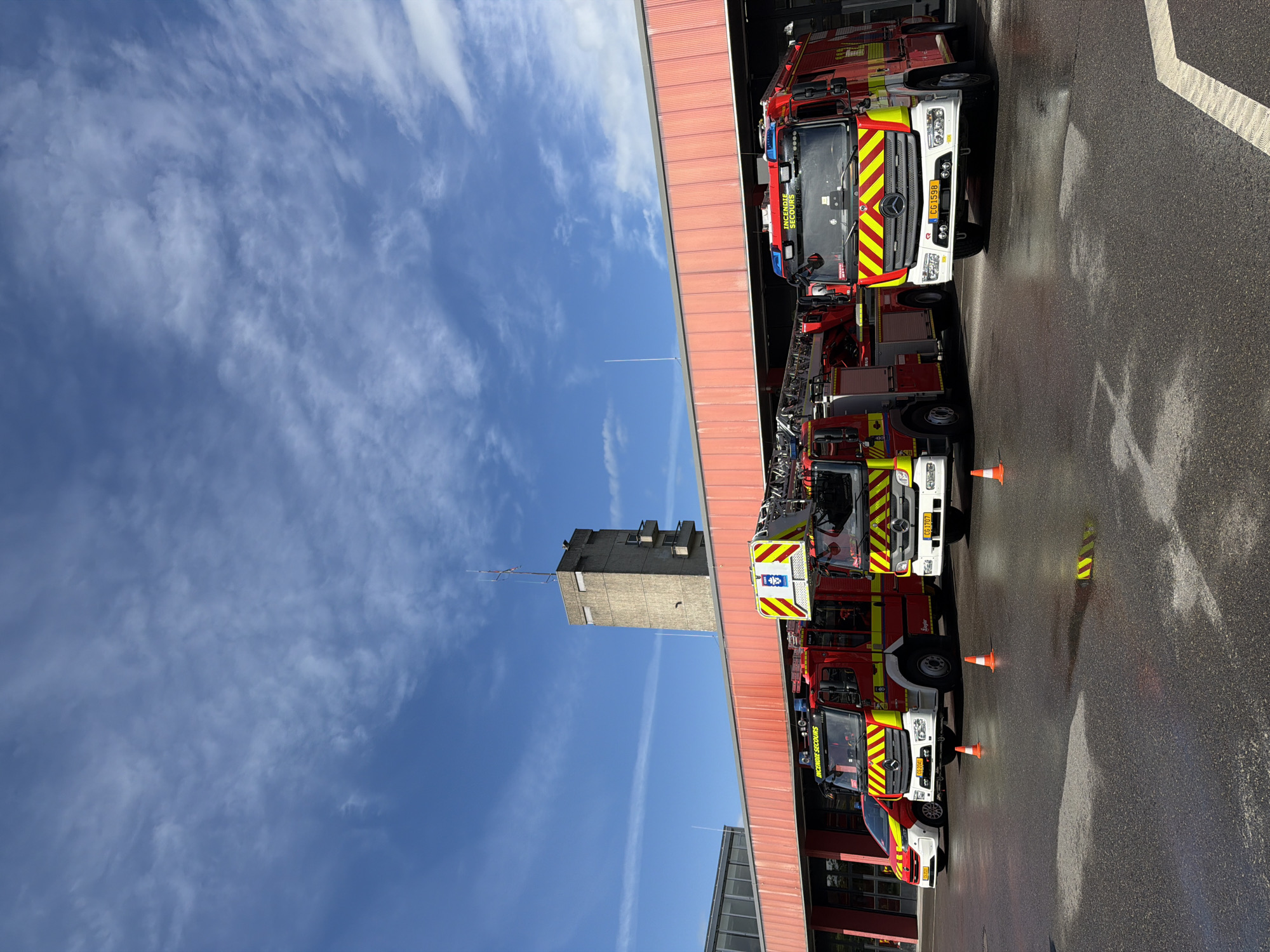 The image shows a fire station in Esch-sur-Alzette, Luxembourg. Four fire trucks are parked in a row in front of the station's bays. Orange traffic cones are placed between the fire trucks. The sky is blue with scattered clouds. A tall concrete tower stands adjacent to the fire station. The ground in front of the station is wet. The license plates of the fire trucks are visible and read "GG 1598," "FCB 7107," and "FCB 9204."