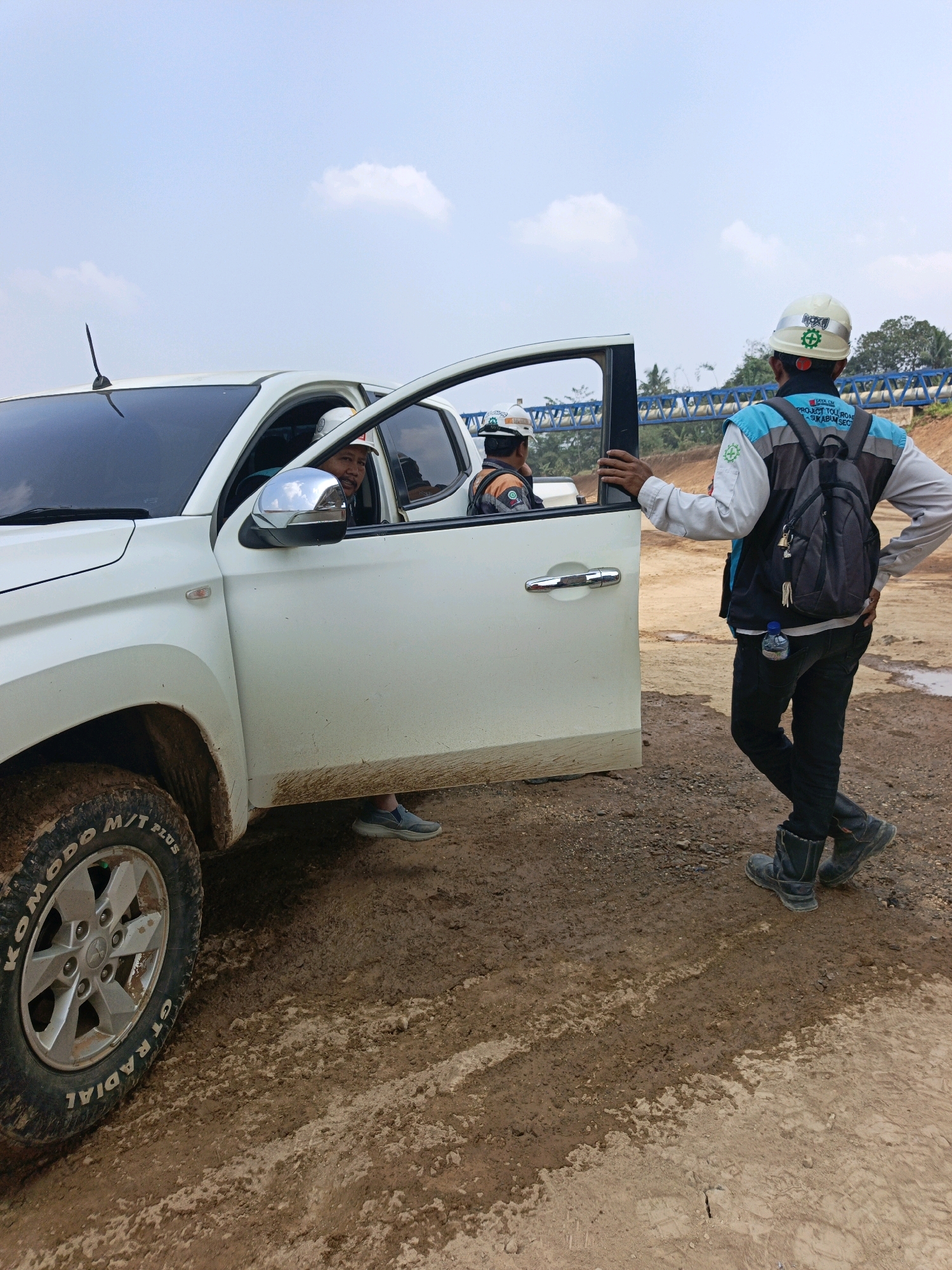 In Cibadak, Indonesia, a white pickup truck is parked on a muddy construction site. The driver, wearing a white hard hat and orange vest, looks out the open driver's side door at a man standing outside. This man, also wearing a white hard hat, a light blue and gray vest, black pants, and black boots, has a backpack on. He is leaning on the open door, facing away from the camera. The truck's tires are covered in mud, and the surrounding ground is wet and muddy, with tire tracks visible. In the background, there is a blue bridge structure and vegetation. The sky is clear with a few scattered clouds.