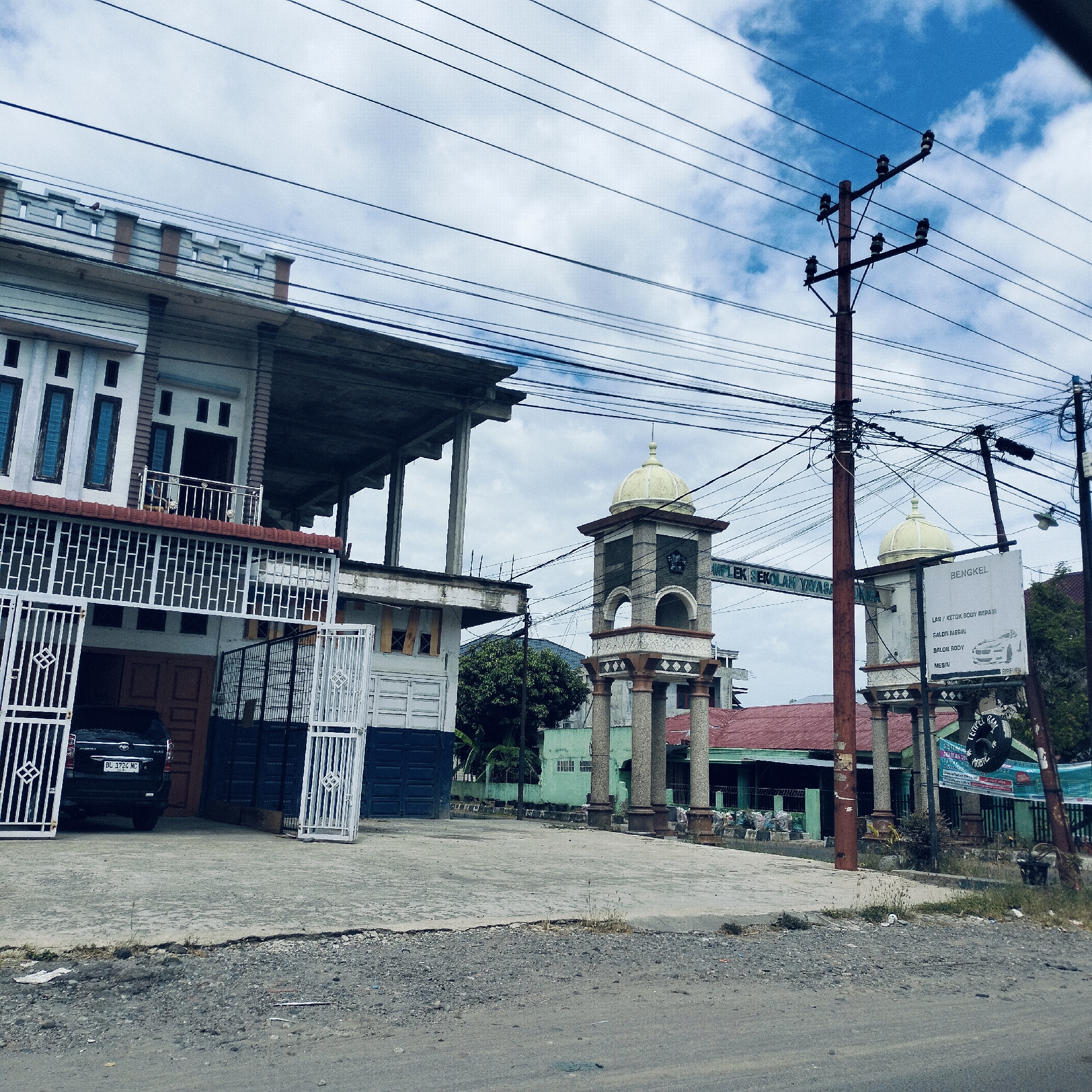 A daytime street view in Cotkeutapang, Indonesia, captures a diverse urban landscape under a partly cloudy sky with prominent blue patches. The scene is dominated by a dense network of overhead power lines crisscrossing the frame, attached to several utility poles.
On the left, a multi-story building, possibly a residence or commercial property, features a white facade with dark-framed windows and an unfinished upper level exposing concrete support structures. White metal gates guard a paved driveway or garage entrance, where a dark-colored car, identifiable by its Indonesian license plate "BL 1724 NC", is partially visible.
In the middle ground, two decorative structures resembling minarets or ornate pillars with small yellow domes stand as an entrance. An overhead sign spans between them, with partial text visible as "MPLEK SEKOLAH YAYASA", likely indicating a "Foundation School Complex". To the right of these structures, a tall utility pole supports a white signboard. The sign, labeled "BENGKEL" (Workshop), lists services such as "LAS / KETOK BODY REPAIR" (Welding / Dent Body Repair), "SALON MESIN" (Engine Wash/Cleaning), "SALON BODY" (Body Wash/Cleaning), and "MESIN" (Engine), accompanied by a graphic of a car and the text "BIREVEN 027". Behind these structures, another building with a red roof and some trees are visible.
The foreground consists of a rough, unpaved road transitioning to a cracked concrete area, suggesting a roadside perspective. No people are visible, and the scene appears to be a typical snapshot of a local street, possibly near a school or religious institution, with commercial activities like an auto repair shop in the vicinity.