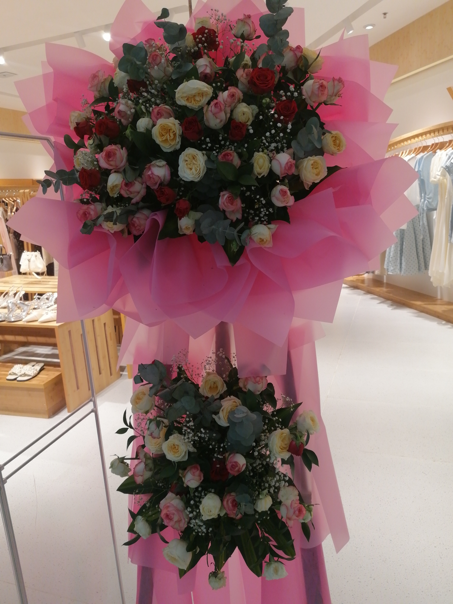 This image captures a meticulously arranged floral display, likely a congratulatory or celebratory bouquet, placed indoors. The bouquet is substantial, featuring a vibrant mix of roses in shades of pink, red, and cream, interspersed with delicate white baby's breath and lush green eucalyptus leaves. The arrangement is dramatically presented, enveloped in layers of translucent pink paper that fans out in a geometric, petal-like design, creating a visually striking effect.
The bouquet is supported by a slender, metallic stand. Below the main arrangement, a second, smaller bouquet echoes the color palette and floral elements of the larger one, also wrapped in pink paper. The backdrop suggests a retail or commercial environment. In the background, racks of clothing are visible, indicating the location might be a boutique or a department store. A wooden display shelf with shoes and bags is also present, further reinforcing the retail setting. The flooring is a pale, speckled surface, and the ceiling features bright, recessed lighting.
While the image provides a detailed view of the floral arrangement and its immediate surroundings, there are no individuals or specific landmarks present that would definitively place it in Da Lat, Vietnam. The scene is generic enough to be found in a commercial establishment in many urban settings.