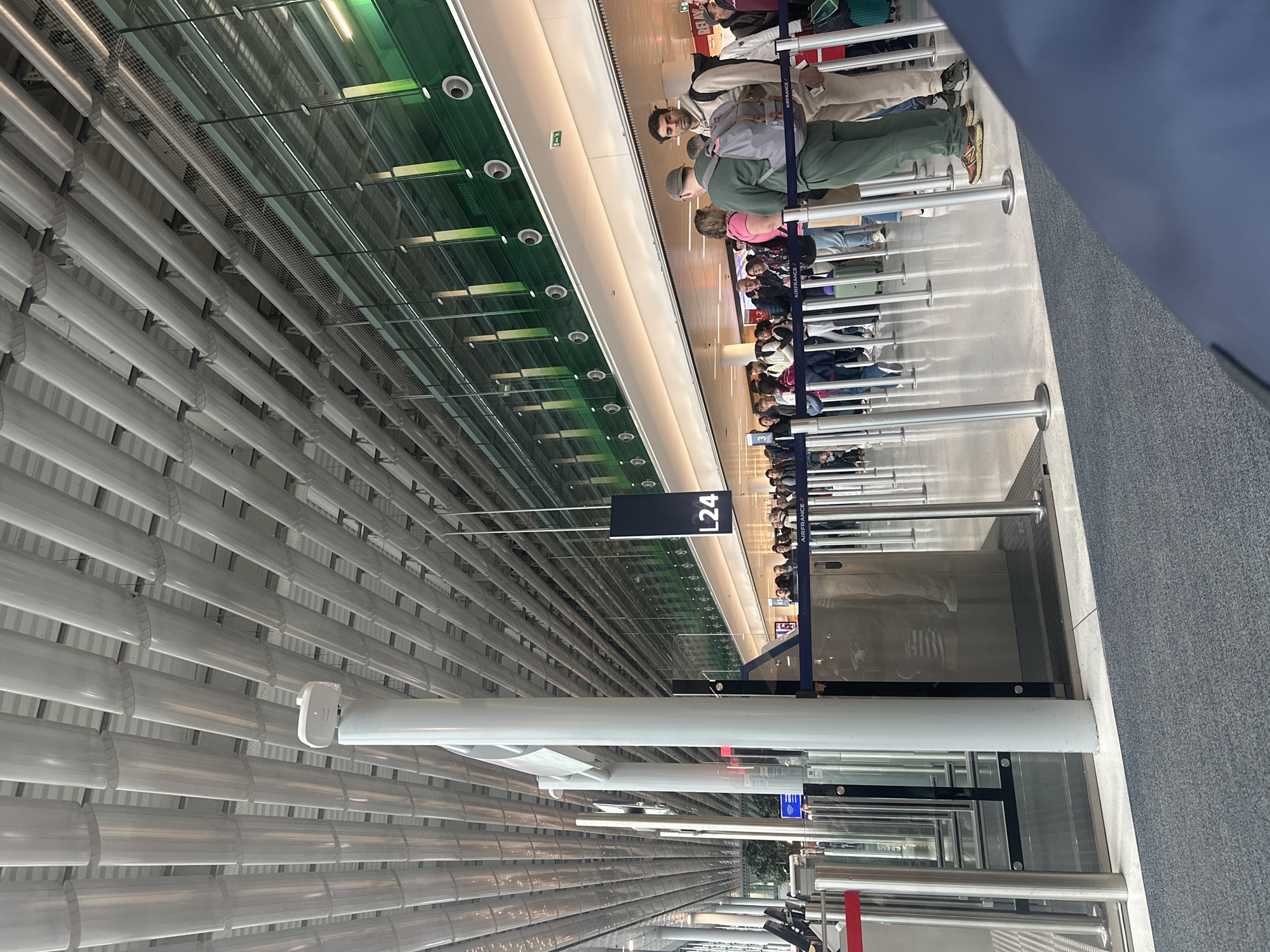 The image depicts a queue of people standing in line at gate L24, inside Charles de Gaulle Airport (CDG) in Le Mesnil-Amelot, France. Passengers are waiting near stanchions. Above them is a white slatted ceiling and green-tinted glass roof, with fluorescent lighting. Many passengers are Air France travelers.