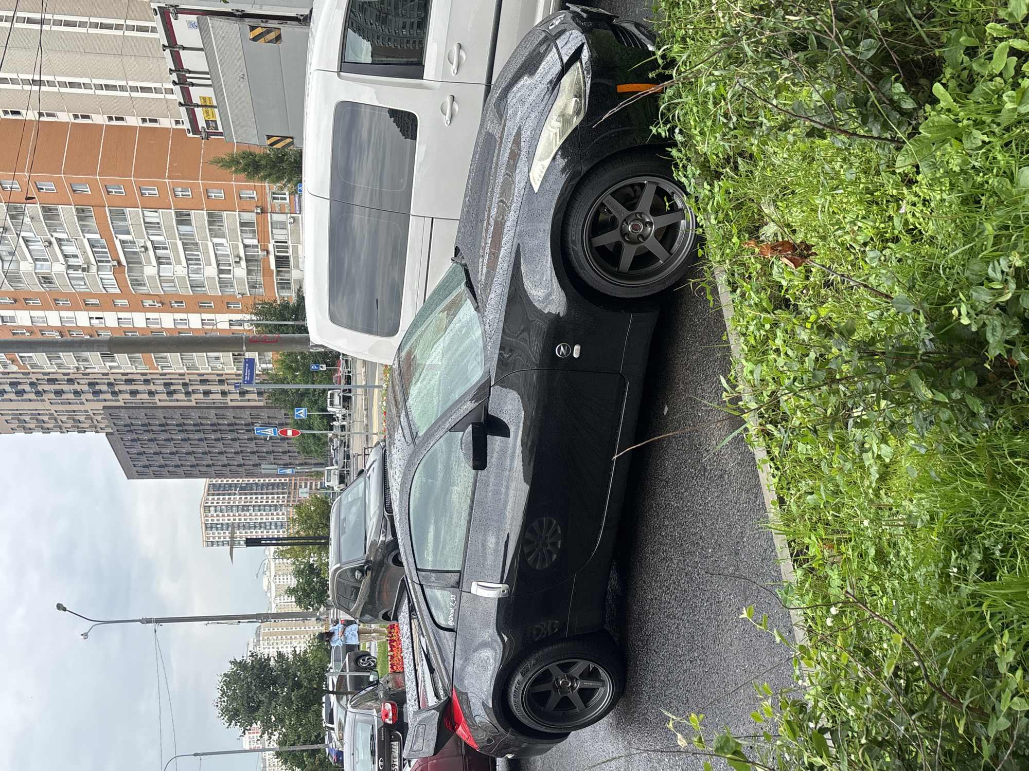 An overcast day in Moskovskiy, Russia, features a black Nissan 350Z parked on a wet asphalt road, likely after recent rain, as evidenced by water droplets on the car and road. The car is positioned to the right of the frame, with its rear spoiler and side profile clearly visible. It has dark gray racing wheels. In the background, several other vehicles, including a white van and other cars, are present. Tall, modern apartment buildings dominate the cityscape, suggesting an urban environment. Dense green foliage, including grass and bushes, lines the foreground, partially obscuring the lower portion of the car. A street lamp is visible in the upper left corner, and a pedestrian signal light with a red circle and a white pedestrian figure is in the distance.