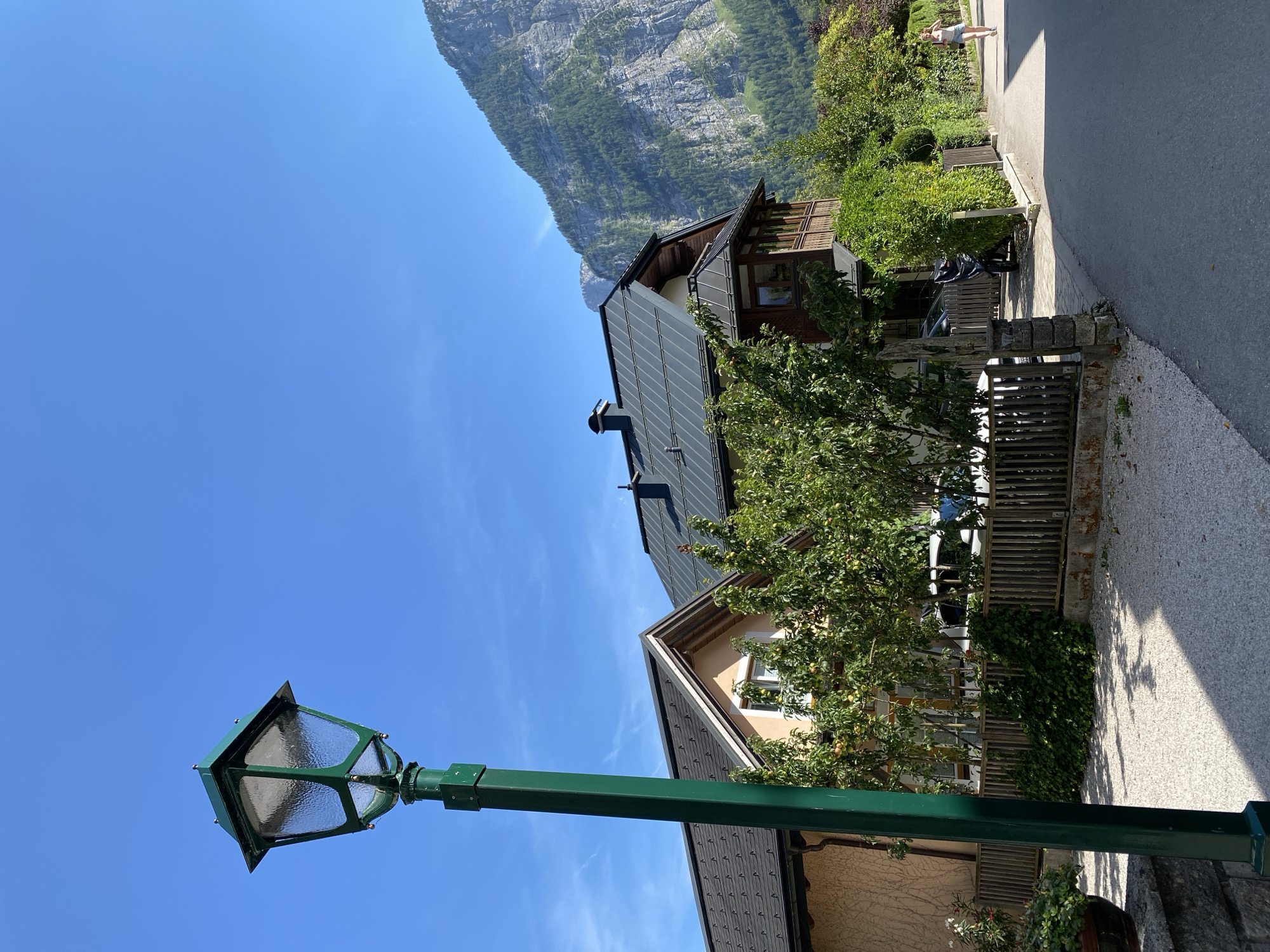 The image captures a sunny daytime scene in Hallstatt, Austria, featuring a street with residential buildings and a mountain backdrop. In the foreground, a green lamppost stands prominently on the left. A paved road runs along the right side of the image, with a pedestrian, a woman in a light-colored top and shorts, walking away from the camera. Adjacent to the road is a wooden fence bordering a gravel driveway and a yard with trees. Behind the fence, houses with dark roofs and light-colored facades are visible. The background is dominated by a steep, rocky mountain covered with scattered green vegetation, under a clear blue sky with faint wispy clouds.
