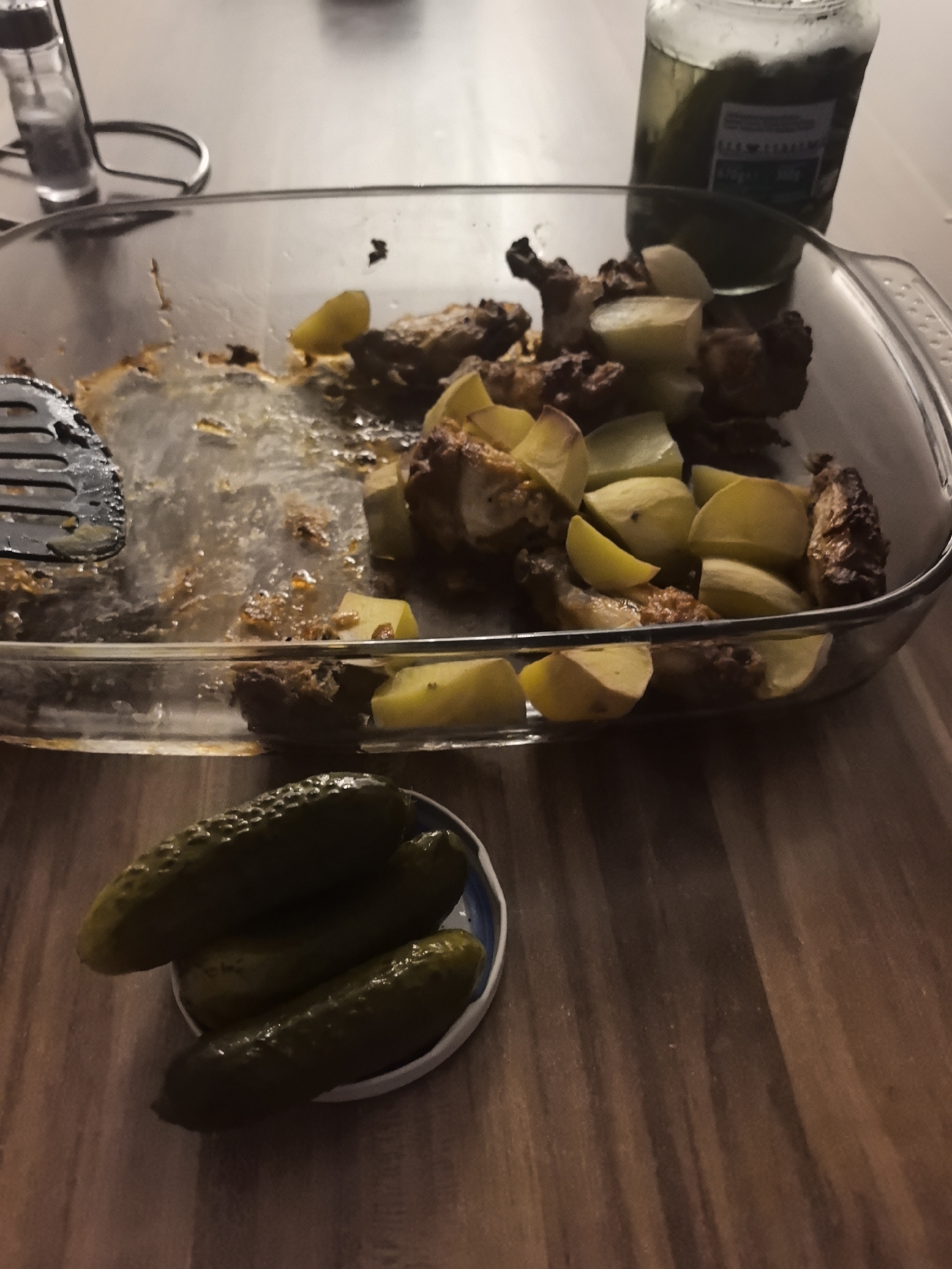 The image depicts a dinner scene, likely indoors, set on a wooden table. In the foreground, there is a small plate containing three pickles. Next to it is a clear glass baking dish, partially filled with leftover food, possibly chicken and potatoes. A black slotted spoon rests within the dish. In the background, a glass jar, presumably containing more pickles, stands upright. A salt shaker sits to the left of the dish. The image was taken at an angle from a high perspective, and it captures a casual, domestic setting, possibly in Hattersheim am Main, Germany.