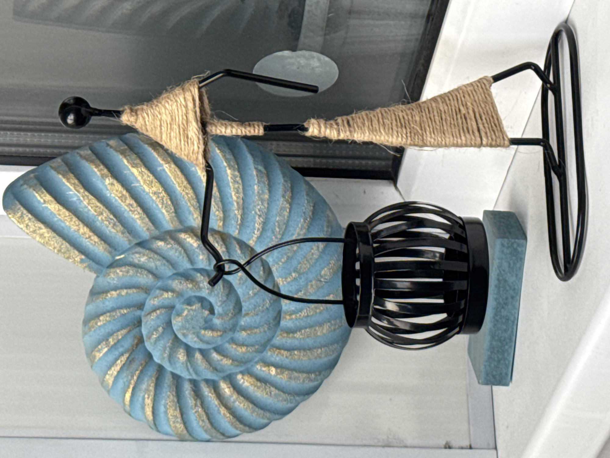 The image depicts a decorative object indoors. The object consists of a stylized metal figurine wrapped with twine, a blue and gold seashell design, and a cylindrical metal and blue base. The figurine appears to be mounted on a wall. The background shows a neutral-colored interior. There is no indication of this image being in Kladovitsy, Russia.