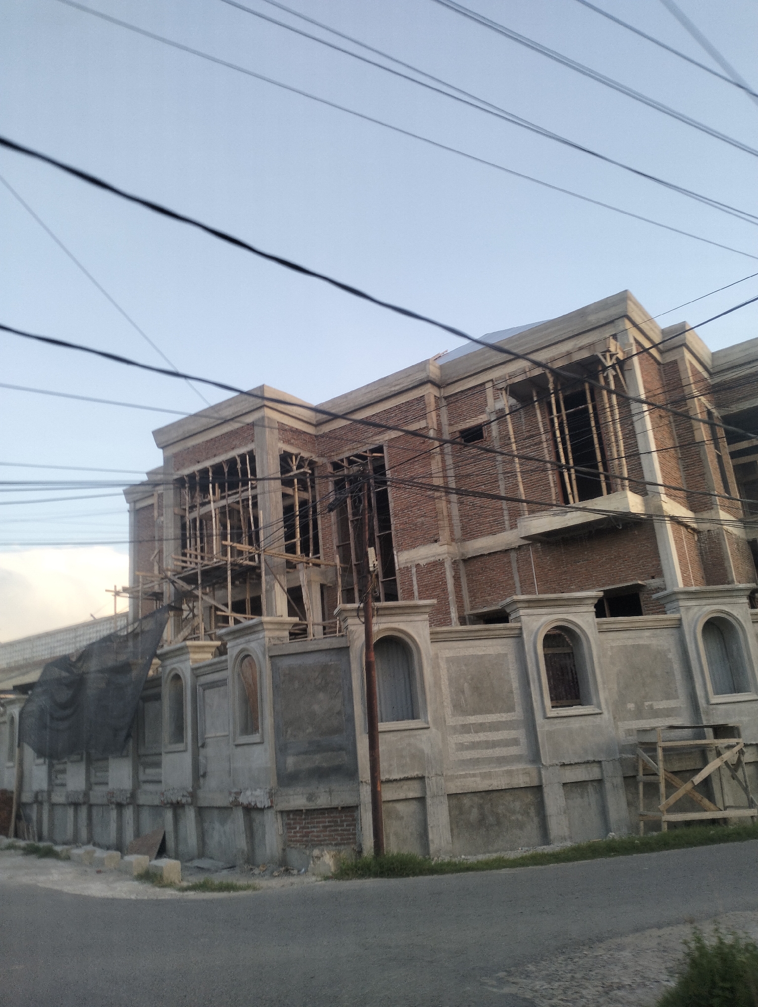 The image shows a multi-story building under construction in Lhokseumawe, Indonesia. The building has a brick facade with concrete trim and decorative elements around the windows and entrance. Scaffolding made of wood is visible on the exterior, indicating ongoing construction work. There are several power lines strung across the sky above the building. The lower part of the building features a decorative concrete wall with arched openings. The ground in front of the building is an asphalt road, and there is some vegetation growing along the roadside. The sky is clear and pale blue, suggesting it might be daytime. No people are visible in the frame. The construction appears to be in an advanced stage, with the main structure mostly in place.