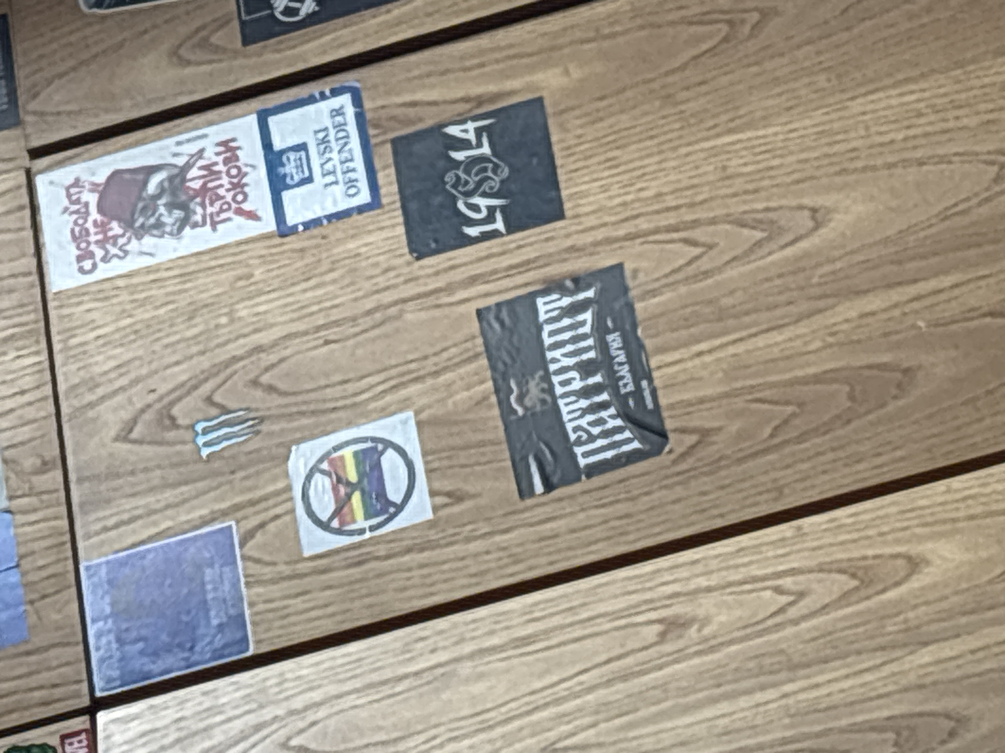 The image displays a collection of seven stickers adhered to a light-brown wooden surface, likely a table or wall panel, characterized by visible wood grain.
One prominent white rectangular sticker features a stylized horned figure with red Cyrillic text on the left that reads "СВОБОДАТА НА ЧЕСТТА Е ТЪРЖИЛ РОКОВИ" (Freedom of Honor is the Triumph of the Fates). On the right side, a blue section contains the English text "LEVSKI OFFENDER" below a crown emblem.
Above and to the right of this sticker is a dark grey/black rectangular sticker with white text "19S14," where the 'S' is stylized as a serpent or dragon. Below it, another dark grey/black sticker reads "ПАТРИОТ" (Patriot) in large white Cyrillic letters, with smaller Cyrillic text, possibly "БЪЛГАРИЯ" (Bulgaria) or "БЪЛГАРИН" (Bulgarian), underneath.
Further down, a small, light blue sticker shows three stylized wavy lines. Adjacent to it, a white circular sticker depicts a symbol resembling a stylized "H" or a cross, filled with rainbow colors (red, yellow, green, blue), which is then overlaid by a red circle with a diagonal line, indicating prohibition or a "no entry" sign. This likely conveys an anti-LGBTQ+ sentiment.
In the bottom-left corner, a faded blue rectangular sticker shows unreadable white text. No specific activity is occurring, and the time of day or weather is not discernible. The stickers suggest themes related to football culture (Levski), nationalism, and social/political views.