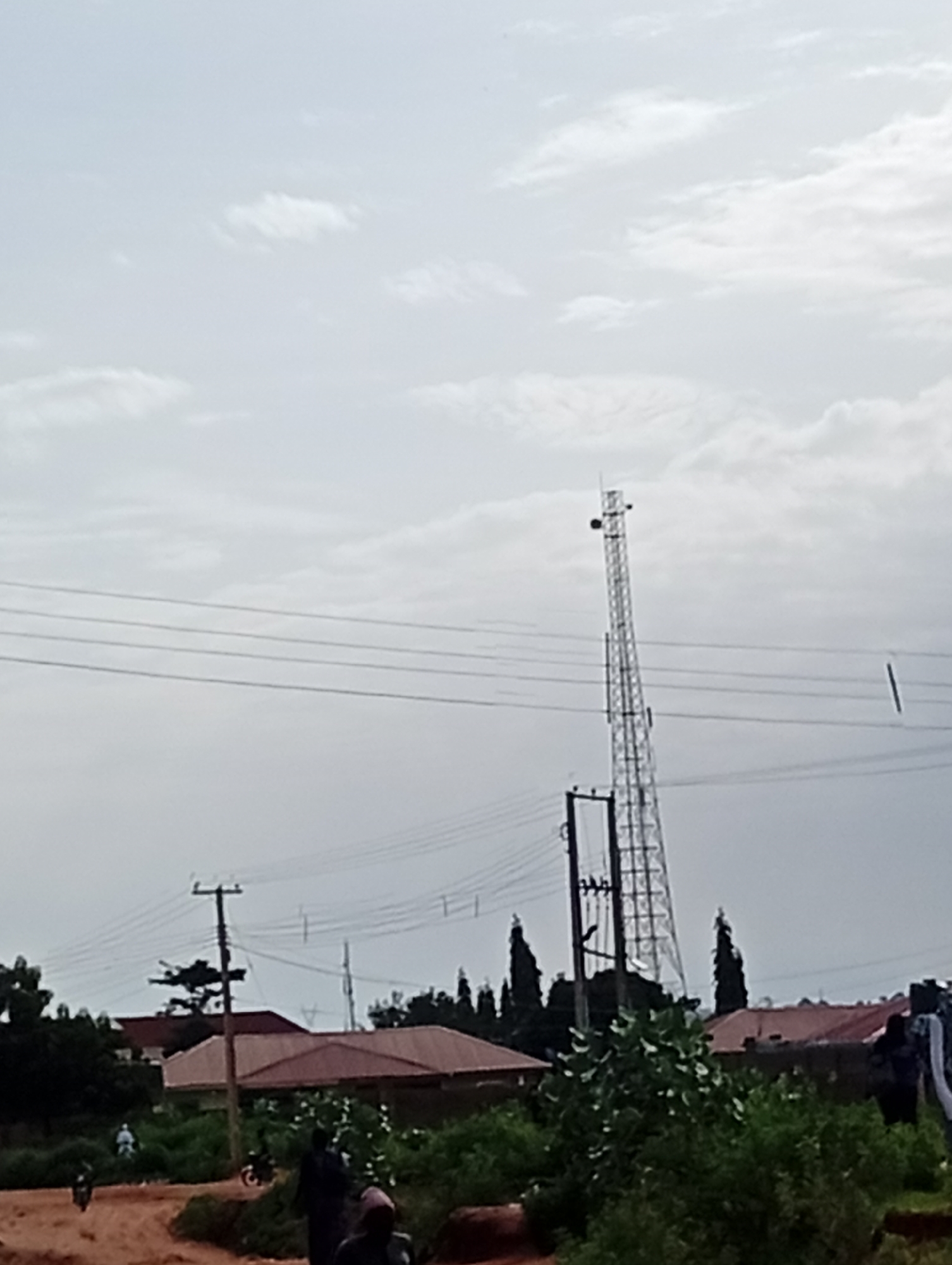 This is an eye-level, medium shot of a rural or semi-urban landscape in Tumfure, Nigeria, under an overcast sky. A tall, lattice telecommunications tower dominates the background, with several power lines strung across the scene. In the foreground and mid-ground, a dirt road runs diagonally, with people and motorcycles present. To the left of the road, there is dense green foliage and trees, with the roofs of a few buildings visible behind them. To the right of the road, more buildings with reddish-brown roofs are seen, along with more vegetation. One individual in dark clothing is walking on the dirt road in the lower right of the frame, and another person on a motorcycle is visible further down the road to the left. The overall impression is one of everyday life in a community with a prominent technological infrastructure.