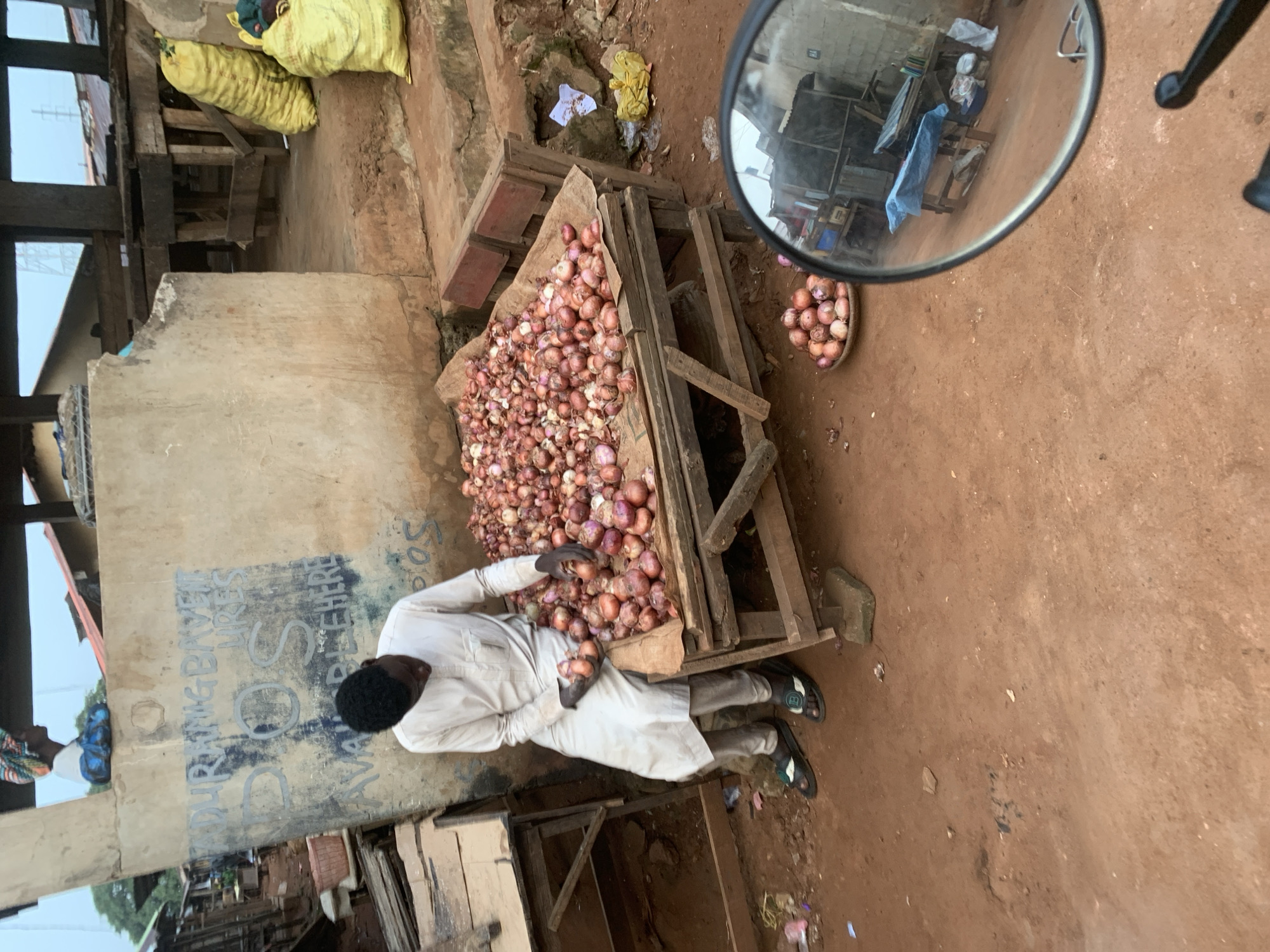 A man dressed in a light-colored traditional outfit sits on the edge of a wooden stall at a market in Shagamu, Nigeria. He is surrounded by a large pile of red onions, and he is holding a few onions in his hands as if examining them. To his left, a concrete wall has faded graffiti on it. Behind him, yellow sacks are stacked, and further back, wooden structures suggest more market stalls or shelters. The ground is made of red earth, and some litter is visible. In the foreground, a motorcycle's rearview mirror reflects a portion of the market scene. Another smaller pile of onions is displayed in a woven basket to the right. The sky appears overcast.