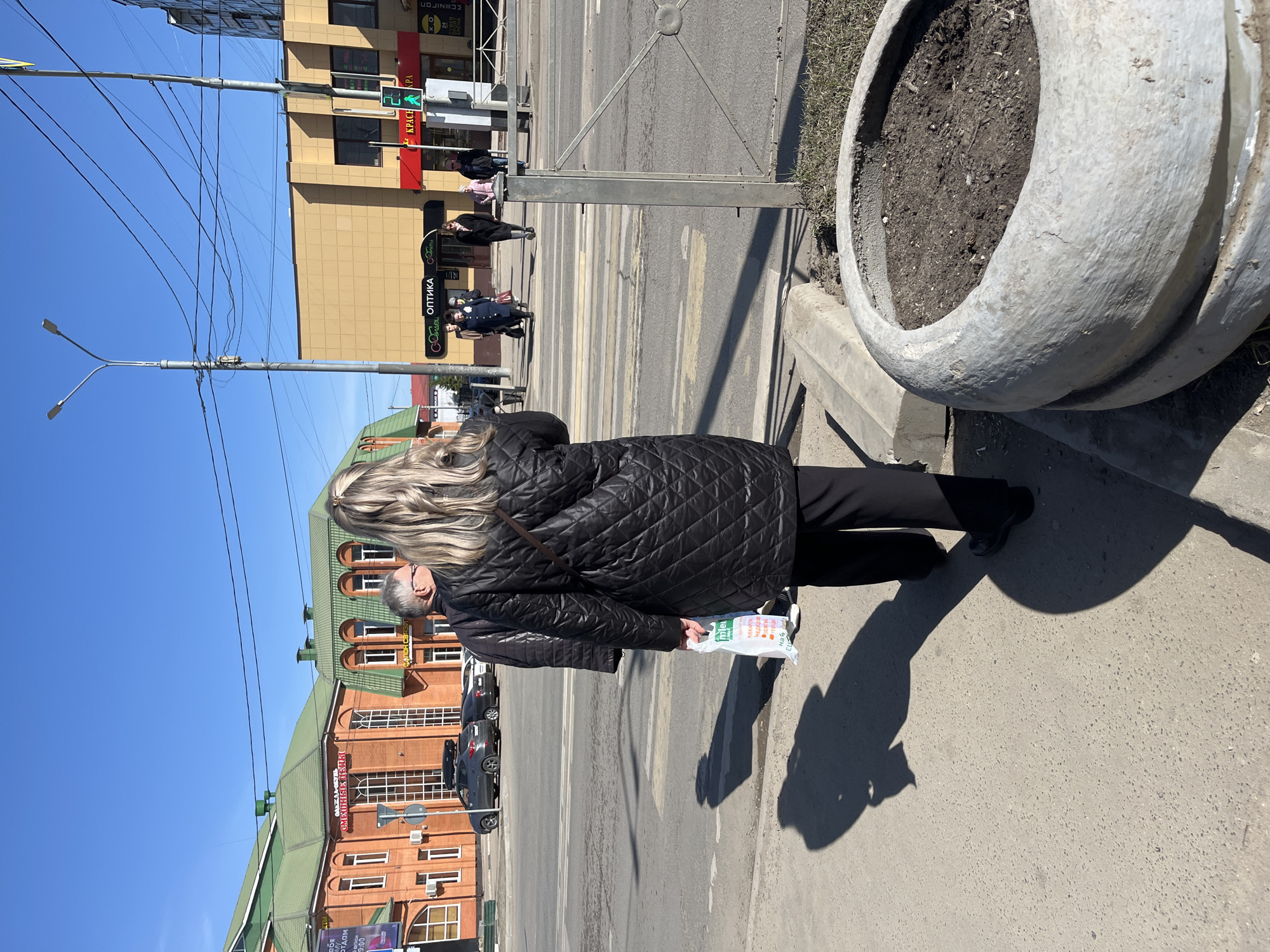 A medium-angle, eye-level outdoor photograph shows a woman in black standing on a sidewalk in what appears to be Klin, Russia.
The woman is facing away from the camera and slightly towards the left. She has blonde hair and is wearing a black quilted jacket and black pants. She holds a white plastic bag in her left hand. Her shadow is visible on the sidewalk to her right.
To the right of the woman is a large, circular, concrete planter with soil. Behind the woman, across the street, is a brick building with green trim. Cars are parked along the street in front of the building. The street itself has a crosswalk with pedestrians crossing. Traffic lights and street signs are also visible.
In the background, across the street from the brick building, is a taller, beige building with several stories. Power lines stretch across the sky. The sky is clear and blue. The general impression is that of a sunny day in an urban environment.
