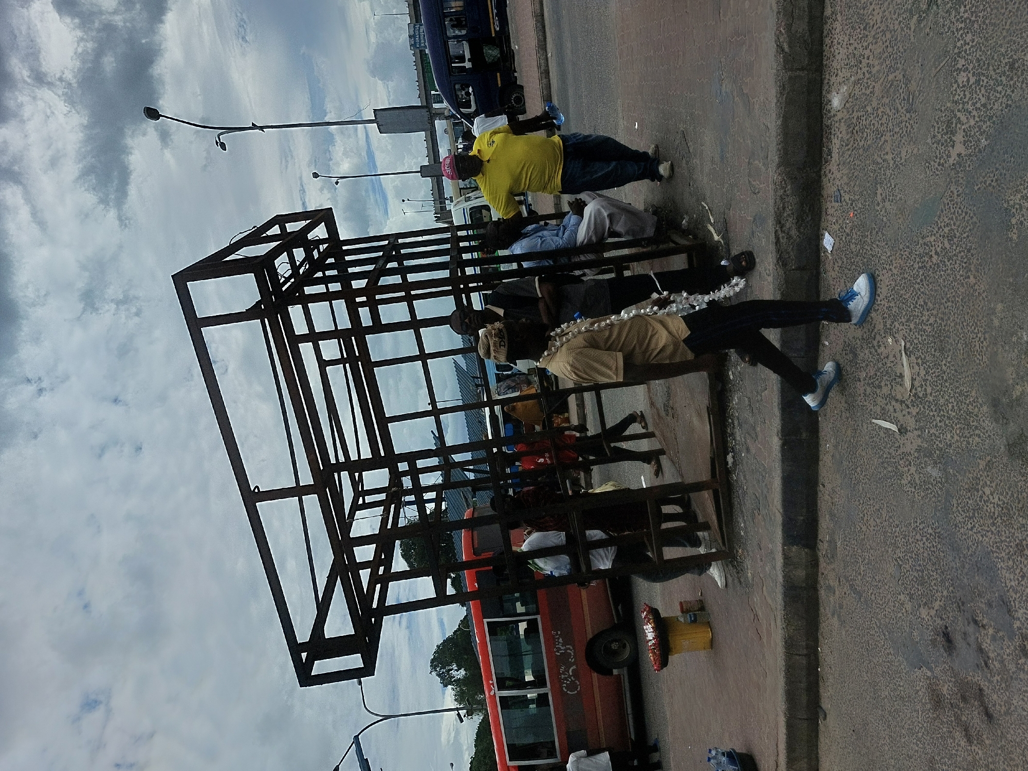 The image depicts an outdoor scene at what appears to be a bus stop in Dar es-Salaam, Tanzania. A metal framework structure stands prominently, possibly part of the bus stop or a nearby construction. Several people are either sitting or standing near the structure and the sidewalk. One person is wearing a yellow shirt. Buses are visible along the road, including one which is orange and another which is blue. The sky is overcast. A street light is visible in the background, as well as other utility poles.