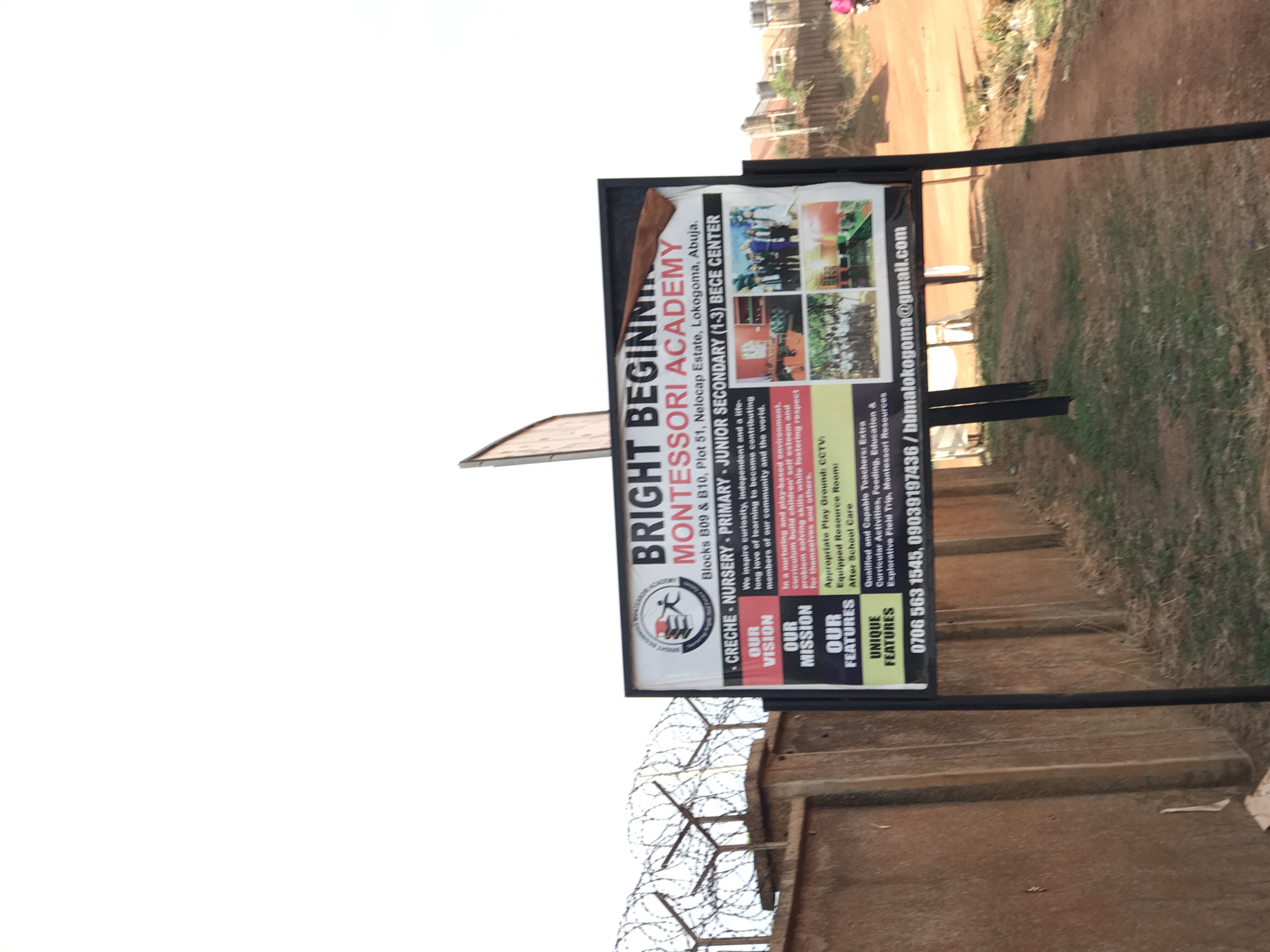 A large outdoor sign for "BRIGHT BEGINNING MONTESSORI ACADEMY" stands upright on a metal frame, positioned beside a dirt path or road. The sign, which appears to be double-sided, faces towards the viewer. The background consists of a clear, bright sky, a dusty ground surface, sparse vegetation, and distant buildings, suggesting a rural or developing urban area. In the lower left foreground, barbed wire atop a wall or fence indicates a property boundary. No people are visible in the immediate outdoor scene.
The sign prominently displays the school's name: "BRIGHT BEGINNING MONTESSORI ACADEMY." Below the name, the address is listed as "Blocks B09 & B10, Plot 51, Nelocap Estate, Lokogoma, Abuja." The educational programs offered include "CRECHE - NURSERY - PRIMARY - JUNIOR SECONDARY (1-3) BECE CENTER." Sections detail the school's "OUR VISION," which aims to inspire curiosity and a lifelong love of learning, and "OUR MISSION," focused on building children's self-esteem and problem-solving skills in a nurturing, play-based environment. "OUR FEATURES" highlight an "Appropriate Play Ground," "CCTV," "Equipped Resource Room," and "After School Care." "UNIQUE FEATURES" list "Qualified and Capable Teachers," "Extra Curricular Activities," "Feeding, Education & Explorative Field Trip," and "Montessori Resources." Contact information includes phone numbers "0706 563 1545, 09039197436" and an email address "bbmalokogoma@gmail.com." Several small images on the sign illustrate children in classrooms, engaging in outdoor play, and participating in what appears to be a school event. The scene is brightly lit, indicative of daytime, under a clear sky.