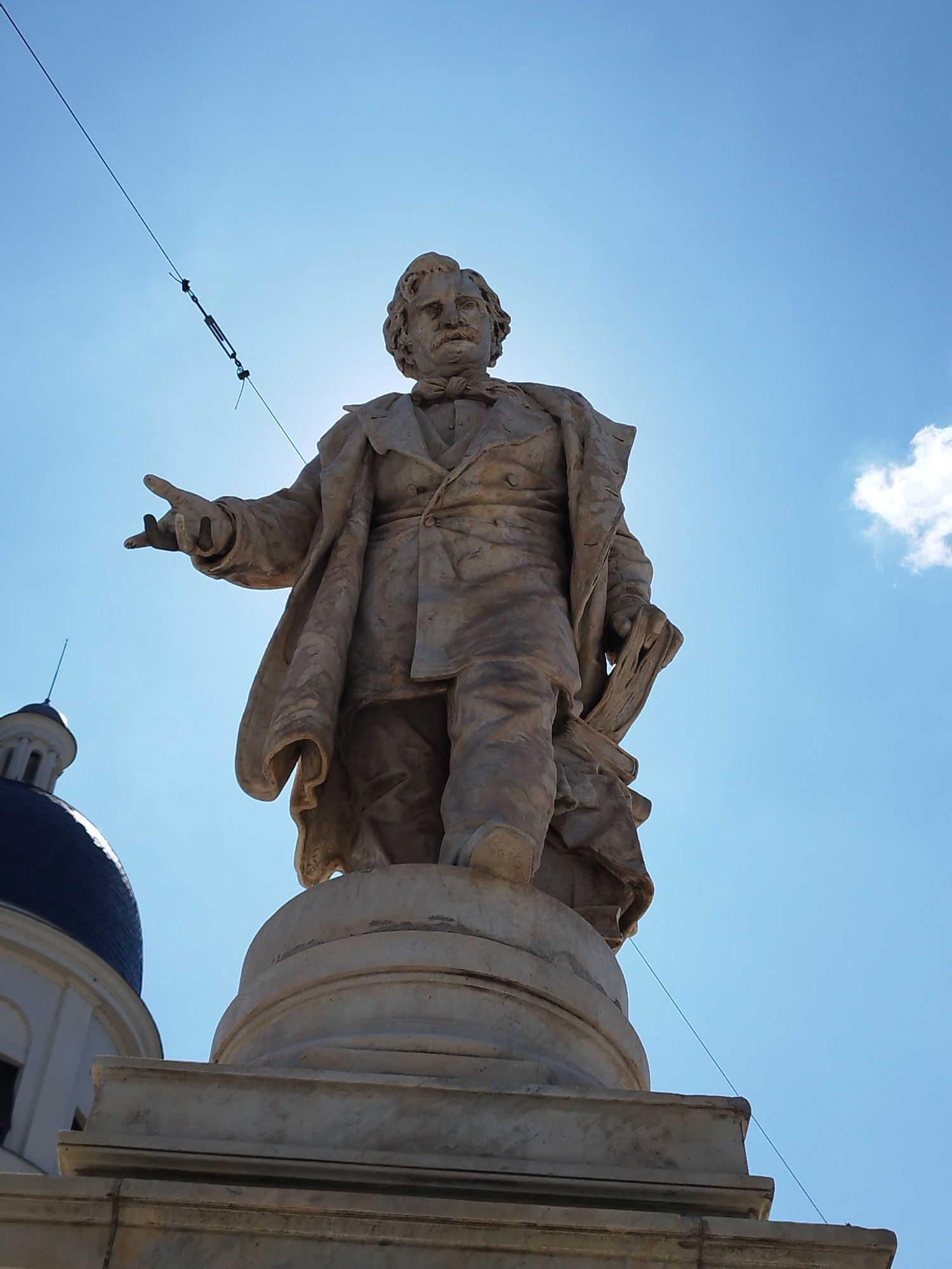 A full shot low angle, eye-level, outdoor, daylight, slightly overcast photograph depicts a marble statue of a man, likely a historical figure, standing on a pedestal in front of a blue sky with a single fluffy white cloud on the right. The statue is made of stone and has a classical appearance. The man is wearing a suit with a cape and appears to be mid-speech or gesturing, with his right arm extended. He is also holding a book in his left hand. The statue is detailed and shows intricate carvings of fabric folds. To the left of the statue, a dome and part of a building are visible, suggesting a public space in a city. The scene is likely located in Bucharest, Romania, given the architectural style which resembles that of buildings found in the Romanian capital. There are two thin, dark wires stretching across the sky, possibly power lines or part of a public infrastructure. The sun is partially obscured by the statue, creating a slight lens flare.