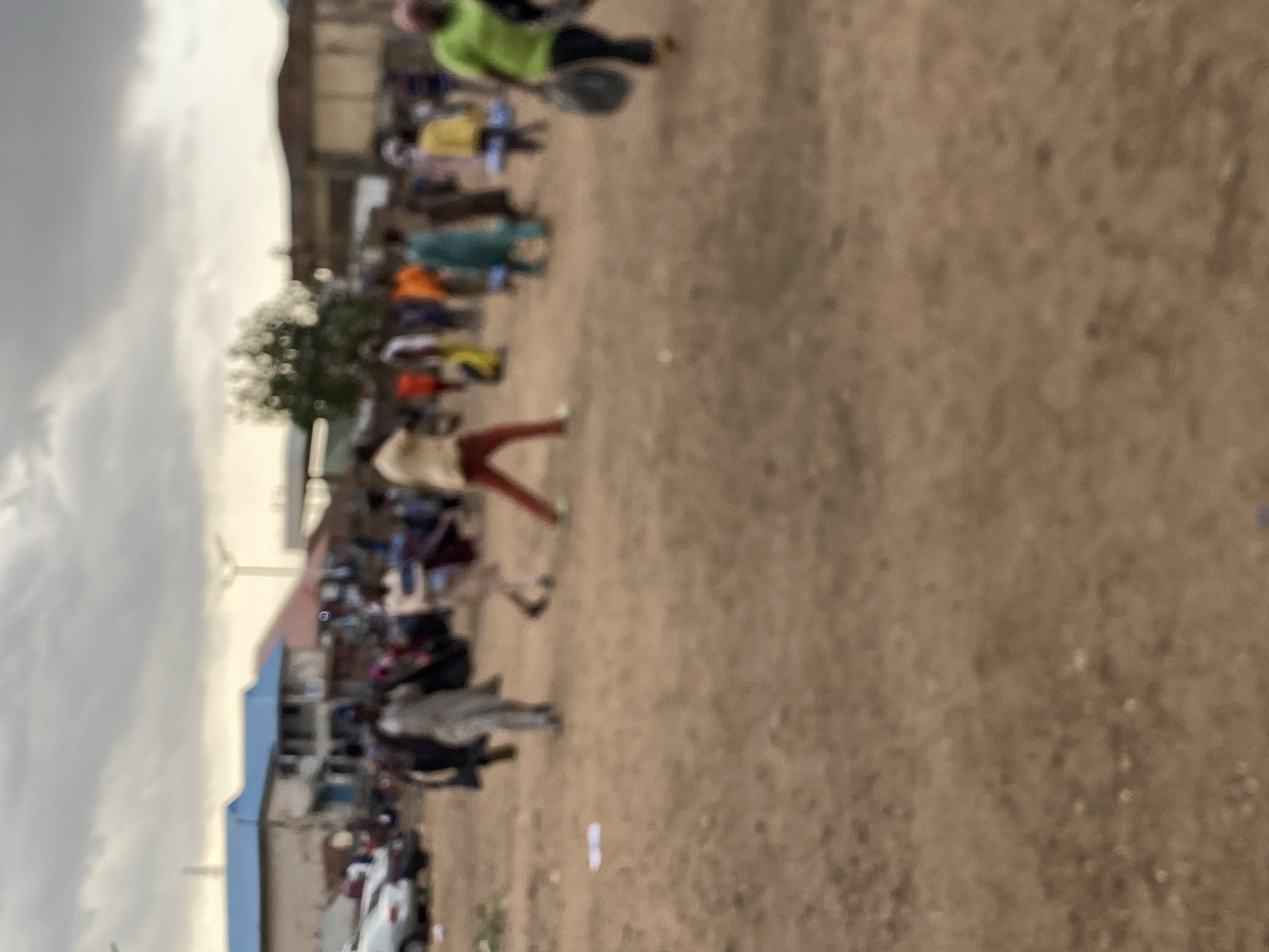 The image depicts an outdoor scene in Bukuru, Nigeria. A group of people are visible, their activities indistinct. Structures, including buildings with blue and white facades, and parked vehicles suggest an urban setting. The ground is dirt. The sky is overcast.