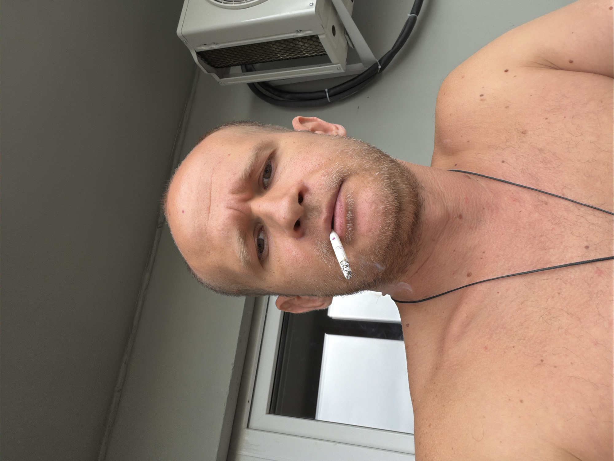 A man is seen from the chest up, shirtless, with a cigarette in his mouth. He has a shaved head and a light beard. Behind him, an air conditioning unit is mounted on a wall. The background suggests an outdoor setting, possibly a balcony or patio, with a glimpse of a window and a gray wall. The image is a close-up selfie. There is no indication of the specific location within Yekaterinburg, Russia.