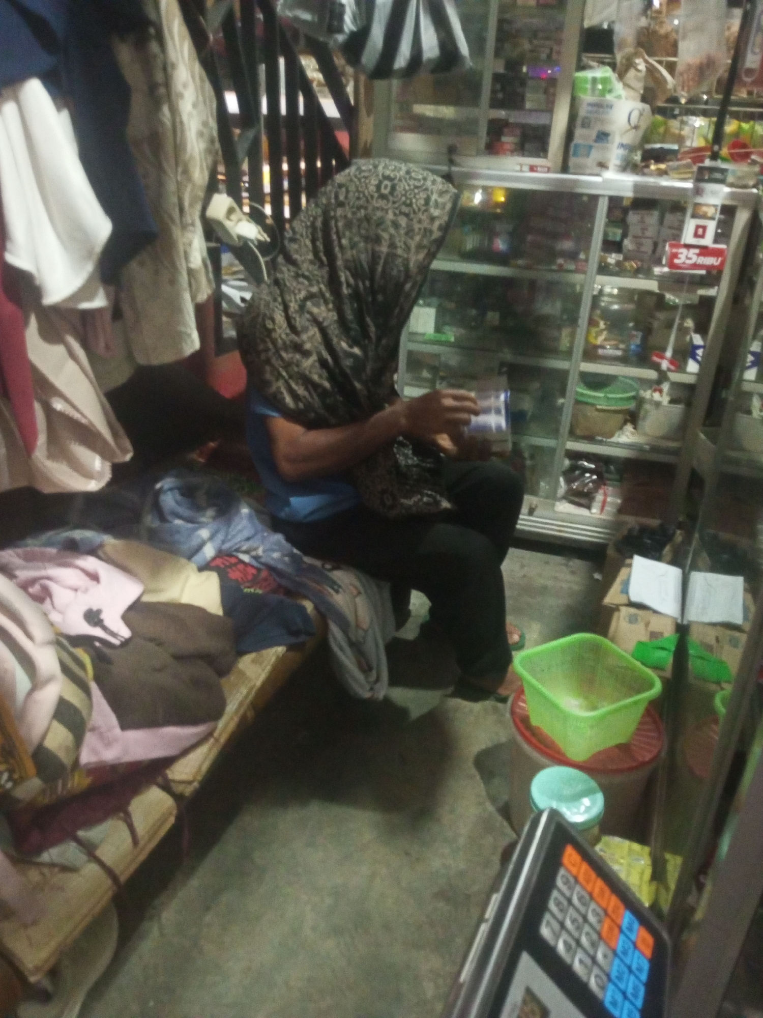 The image depicts an interior scene, likely a shop or stall, with a cluttered arrangement of goods. In the foreground, a person wearing a dark headscarf and a blue shirt is seated on a low wooden platform covered with blankets and clothing. They are holding a clear plastic container with a blue lid and appear to be interacting with its contents. To the left of the seated person, various garments are draped over a rack. Behind them, a glass display cabinet is filled with packaged items and products, some with visible labels, including one that reads "35 RIBU". Several cardboard boxes and a green plastic basin are also visible in the environment. The lighting is dim, suggesting an indoor setting at night or in a poorly lit area. The overall impression is one of a humble, possibly informal, retail space. The location is identified as Andongsili, Indonesia.