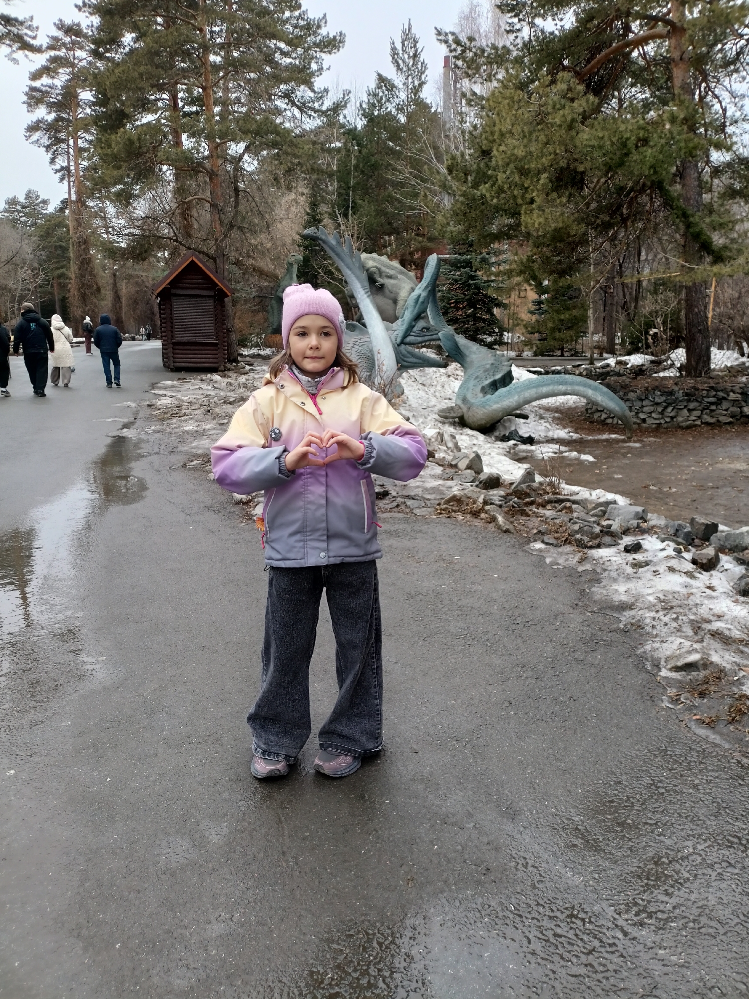A young girl stands on a paved walkway. She wears a pink hat, a yellow and grey jacket, and dark jeans. She is making a heart shape with her hands. Behind her, there is a sculpture that resembles a dinosaur skeleton. To the left, people are walking away from the camera. A small wooden structure is also visible. The ground is wet, and there are patches of snow near a rocky area and water. The scene is outdoors, with trees in the background. The image was captured in the city of Novosibirsk, Russia.