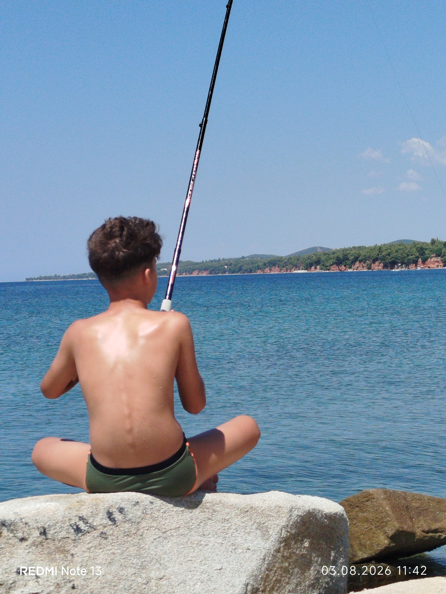 A young boy, seen from behind, sits cross-legged on large rocks by the sea, holding a fishing rod. He is wearing green swimming trunks and his back is glistening, suggesting he might be wet or has been in the sun. The boy appears to be engaged in fishing.
The scene is set against a bright blue sky and a calm, blue sea. In the distance, a tree-covered coastline with red cliffs is visible. The water is tranquil with gentle ripples. The lighting suggests it is daytime, likely midday or early afternoon, with bright sunshine.
The image was taken in the Sithonia Municipal Unit, Greece. Visible text at the bottom left of the image reads "REDMI Note 13", and at the bottom right, it reads "03.08.2026 11:42", indicating the date and time the photo was captured. The overall mood of the image is peaceful and serene, capturing a moment of quiet activity in a beautiful coastal environment.