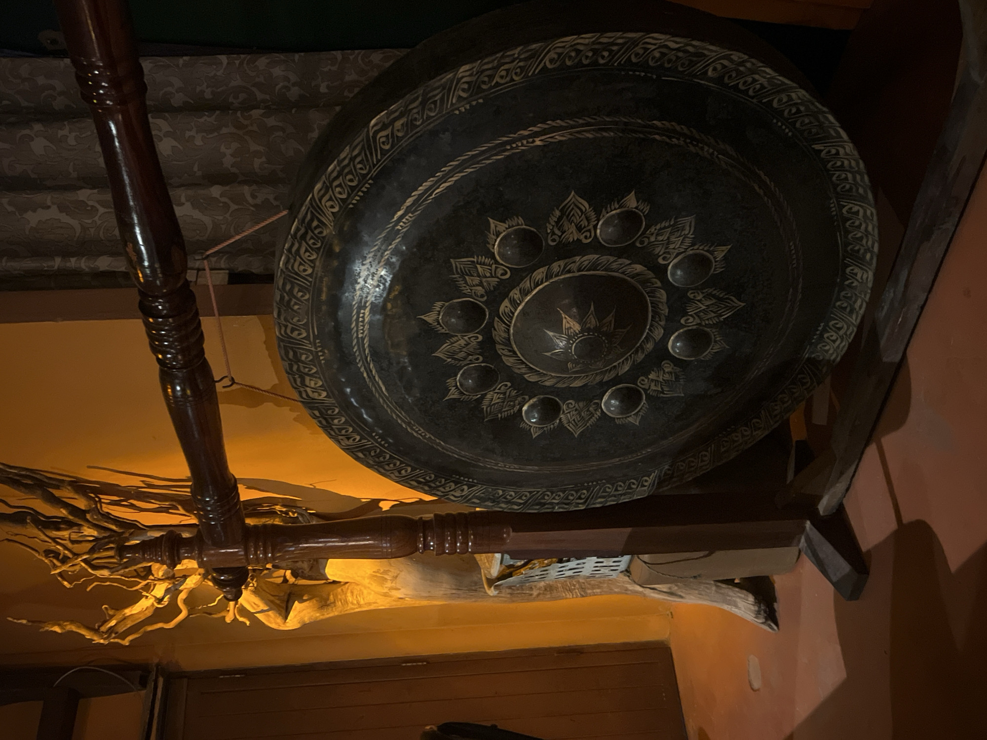The image, taken indoors in Baan Lamai, Thailand, features a large, ornate gong hanging from the ceiling. The gong, round and dark, is decorated with a circular pattern of raised bumps and floral or leaf-like designs. It is suspended by a cord or rope. To the left of the gong is a dark, wooden, intricately carved pole, likely a part of the hanging structure. Underneath the gong and the pole, a decorative arrangement of branches or driftwood is visible. The walls are painted in two different colors, with a bright orange/yellow on the left and a reddish-brown on the right. A dark wooden door or panel is at the bottom center of the frame. The ceiling above the gong has a textured, patterned covering.