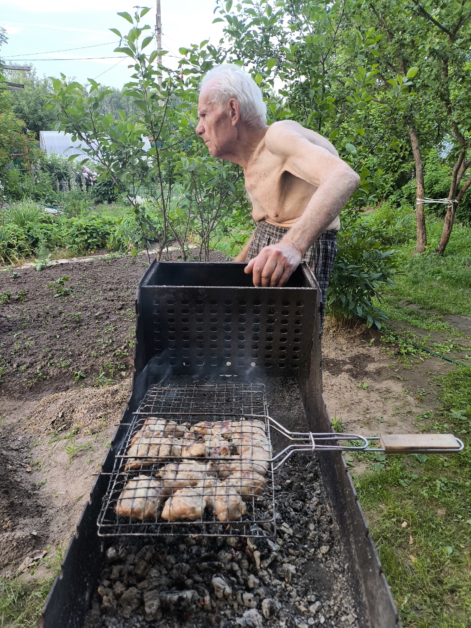 In an outdoor, possibly rural setting, an elderly, shirtless man with white hair stands beside a charcoal grill. He is wearing plaid shorts and his hands are positioned over the top of the grill. The grill contains a wire rack with several pieces of cooked meat, emitting smoke. The background shows lush green trees, plants, and a dirt path. The overall scene depicts a man barbecuing outdoors.