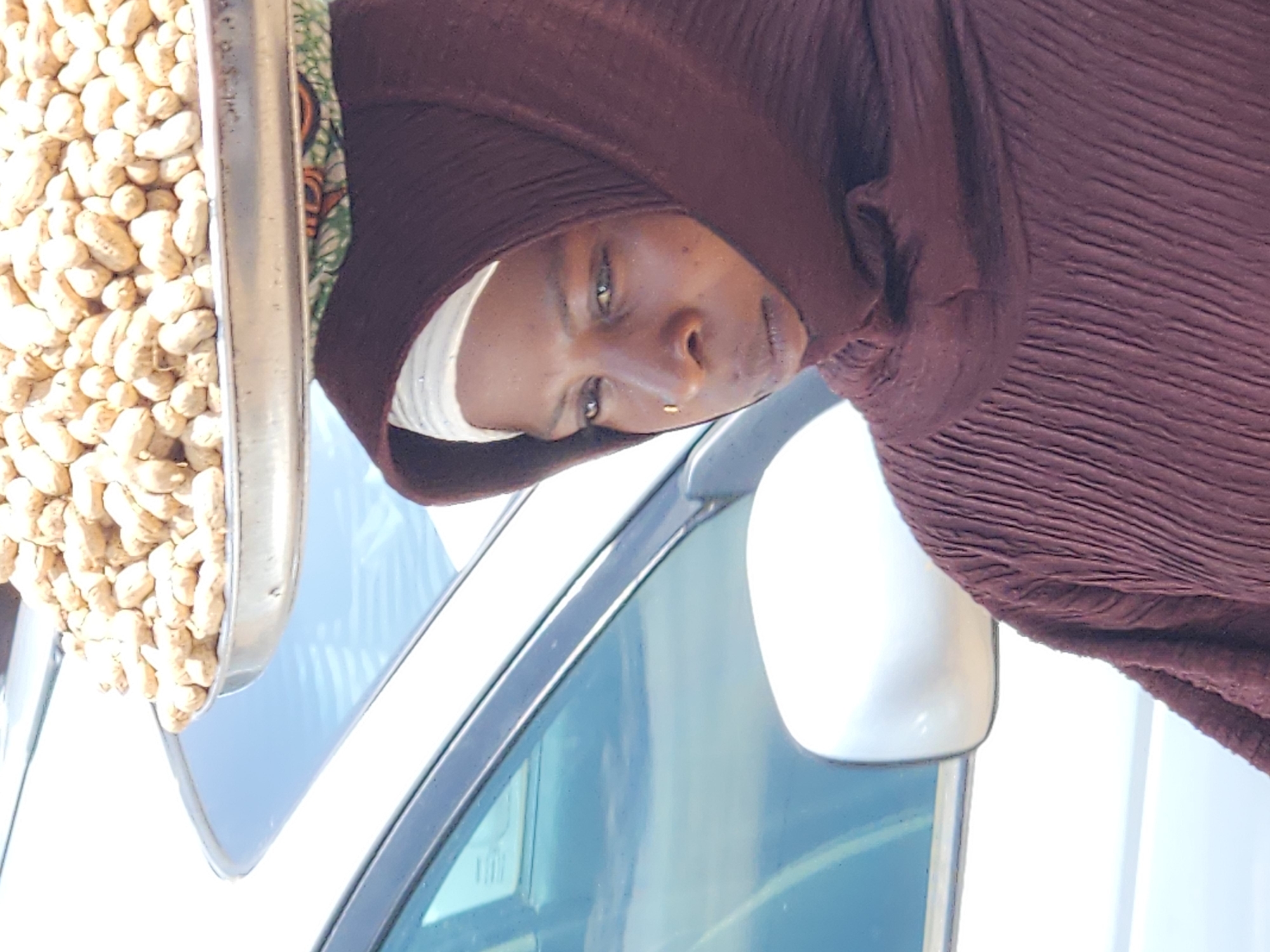 A woman in Jos, Nigeria, is pictured with a tray of peanuts balanced on her head. She wears a brown head covering and a matching brown top with a pleated texture. A gold nose ring is visible on her right nostril. The background consists of the side of a white car, with its side mirror and part of the windshield visible. The lighting suggests it is daytime.