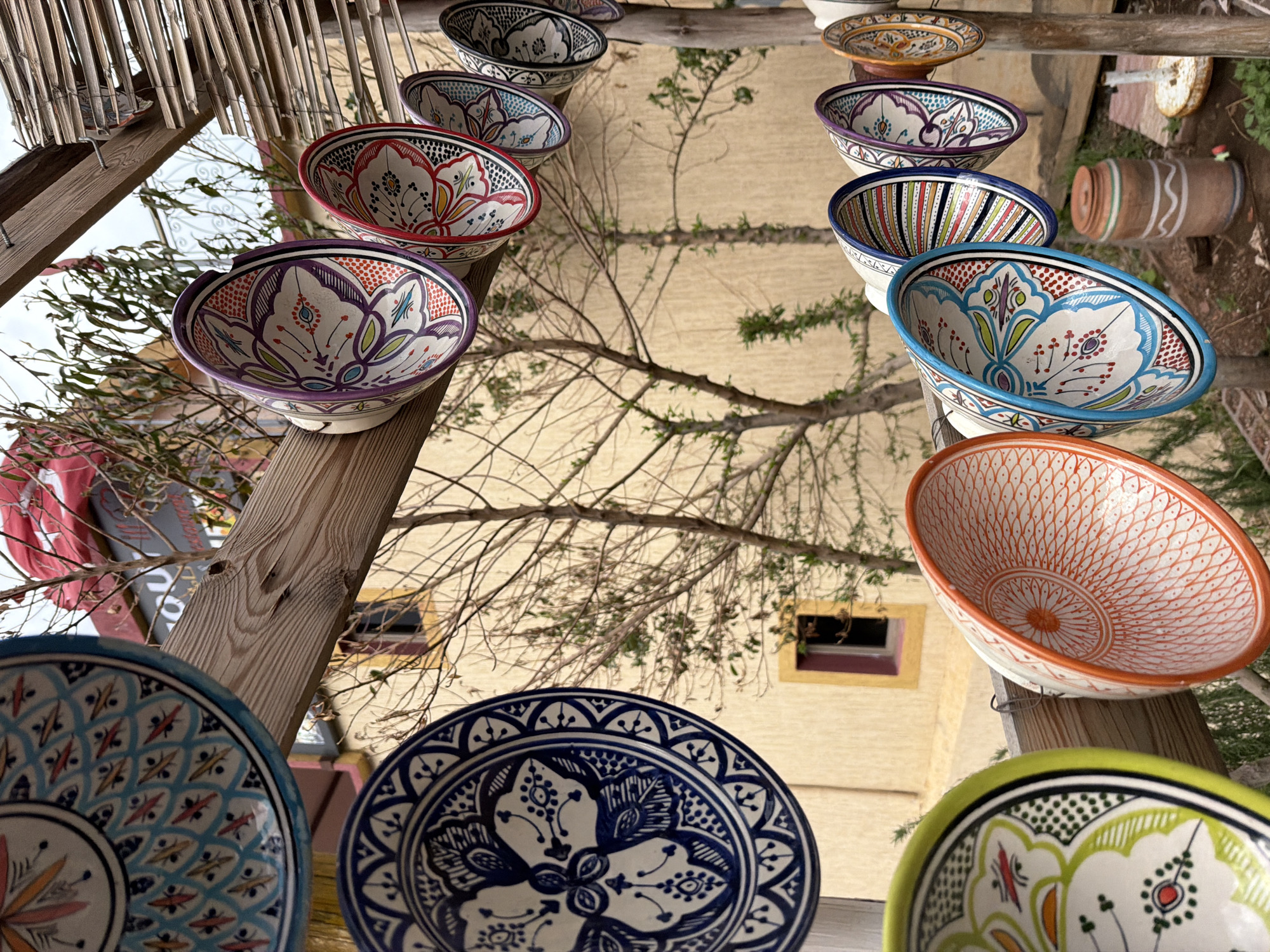 The image depicts an outdoor display of Moroccan pottery in Zerkten, Morocco. The pottery, a mix of bowls and plates, is arranged on a wooden structure. The items showcase traditional Moroccan designs with vibrant colors. In the background, a light-colored building with a window is visible, partially obscured by tree branches. Other elements include a reed roof structure.