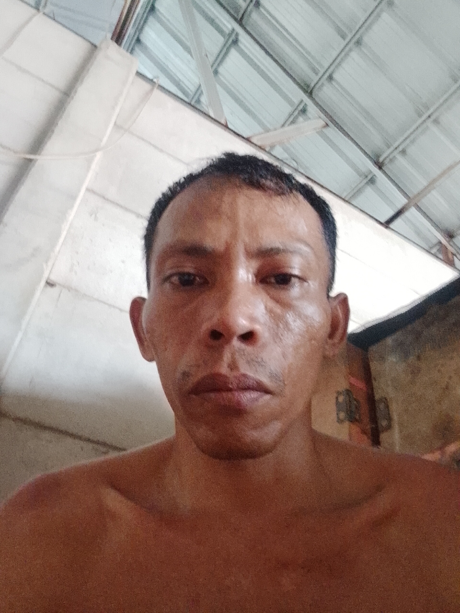 A close-up, slightly upward-angled photograph shows a shirtless man with short, dark hair and a mustache. His skin appears slightly wet or oily. The background features a ceiling with a mix of white painted surfaces and corrugated metal. The photograph was taken indoors, likely in a residential setting in Bukit Ketok, Indonesia, as specified. Details such as the lack of a defined background and the close proximity to the subject suggest the photo may be a selfie.