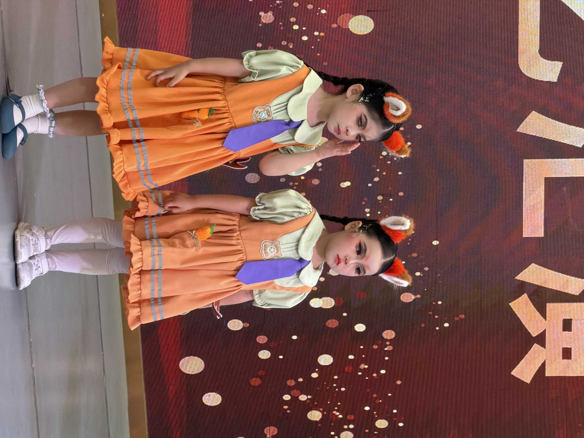 Two young girls are standing on a stage in front of a large screen displaying large characters. The girls are dressed in matching orange dresses with light green collars and purple ties. They are also wearing fox ears on headbands. The girl on the left is touching her face, and the girl on the right is looking forward. The stage floor is a light gray color, and there is a wooden or golden border along the bottom of the screen. The screen has a reddish hue and appears to be a digital display with some blurred lights scattered across it. This scene is likely from a performance or event taking place in the Jiulongpo District of Chongqing, China.
