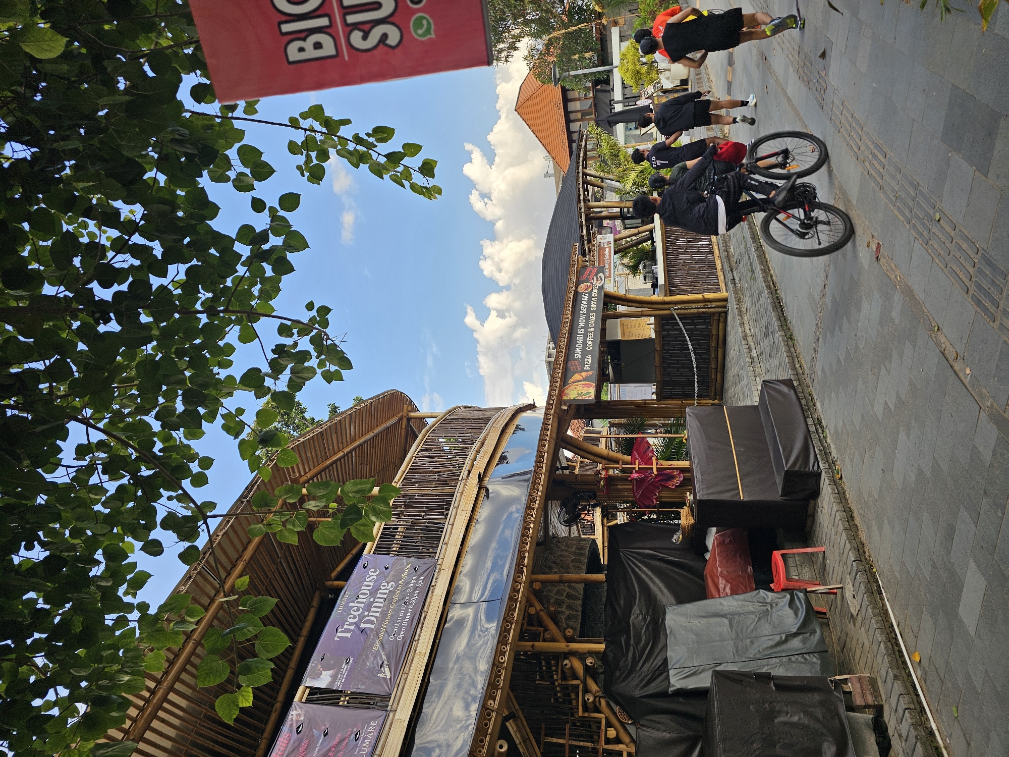 This image captures a street scene in Denpasar, Indonesia, under a bright blue sky with scattered clouds. The focal point is a two-story bamboo structure, likely a restaurant or dining establishment, with a balcony railing and signage that reads "Treehouse Dining." In front of the building, black tarps cover various objects, and a small red stool is visible.
To the right of the bamboo structure, a row of food stalls with bamboo accents is present, displaying menus and offering various food and beverage options. A sign on one stall advertises "SUNDARI IS 'NOW' SERVING PIZZA, COFFEE & CAKES."
Several people are engaged in activities on the paved walkway. A person is riding a bicycle, while others are jogging. The individuals are dressed in casual sportswear, with some wearing black hoodies and shorts, and others in athletic attire. The environment appears to be an outdoor public space, possibly a promenade or pedestrian area adjacent to the establishments. The presence of lush green foliage overhead suggests a tropical setting. A large red banner with white text, partially visible on the right side of the frame, includes the words "BIG" and "SU," possibly indicating another business or promotion.