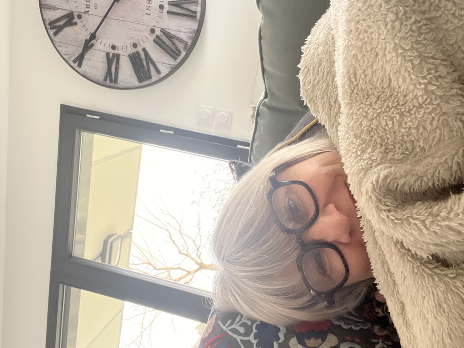 A woman with light grey hair and dark-framed glasses is partially visible, reclining on a dark green sofa, covered from the nose down by a light beige, textured blanket. Her head rests on a patterned pillow. In the background, a large window with dark frames reveals a bare-branched tree and a light-colored outdoor umbrella or awning structure against a bright sky. To the right of the window, on a white wall, a large circular wall clock with Roman numerals and a distressed wooden appearance is mounted. The clock features text, including "Antiquité" and "1880." Below the clock, two white electrical outlets are visible. The scene is located in Chambœuf, France.