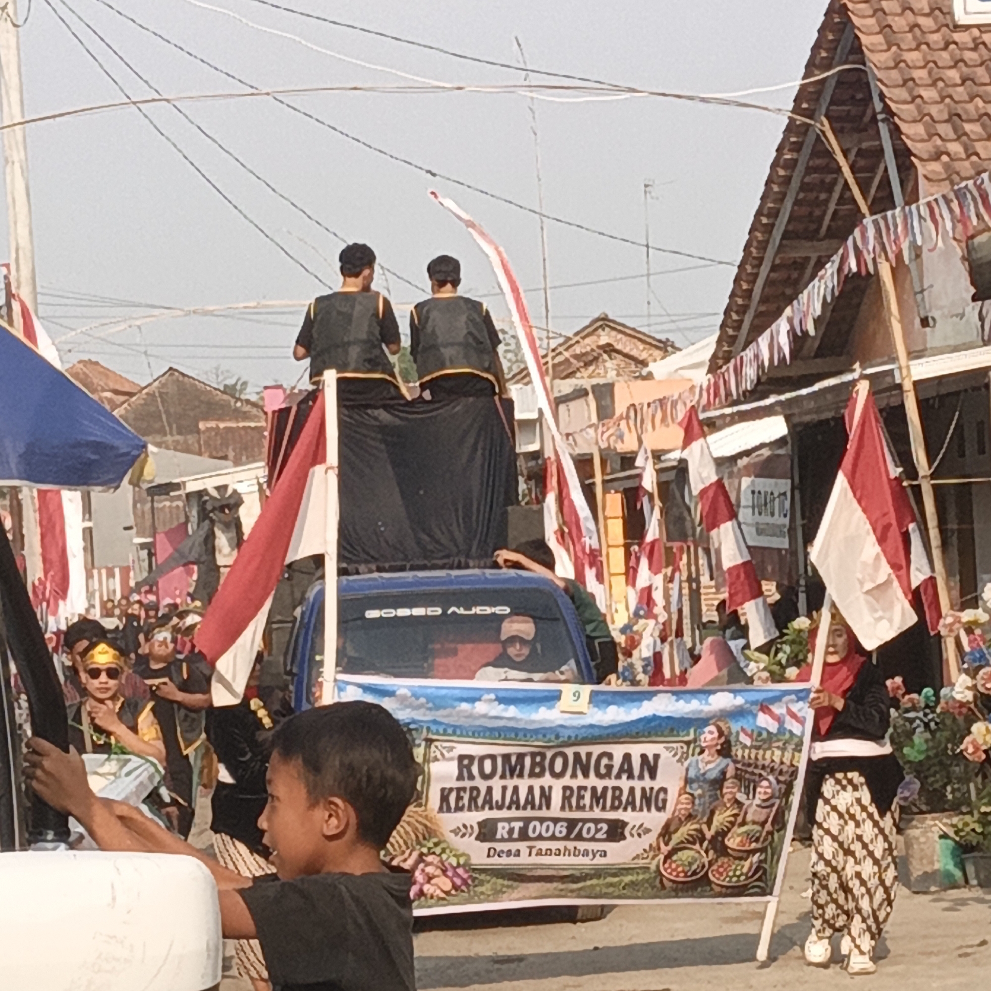 The image depicts a street scene in Tanahbaya, Indonesia, likely during a parade or cultural event. In the foreground, a young boy in a black t-shirt faces to the right, holding a black pole. Behind him, a blue truck is decorated with a banner that reads "ROMBONGAN KERAJAAN REMBANG RT 006/02 Desa Tanahbaya," featuring images of people and Indonesian flags. Two individuals in black vests and head coverings stand on top of the truck. Several Indonesian flags, red and white, are prominently displayed, along with festive red and white bunting hanging from buildings. A group of people are visible in the background, some wearing traditional attire and headwear. The environment appears to be a residential street with buildings on either side. The sky is bright, suggesting daytime.