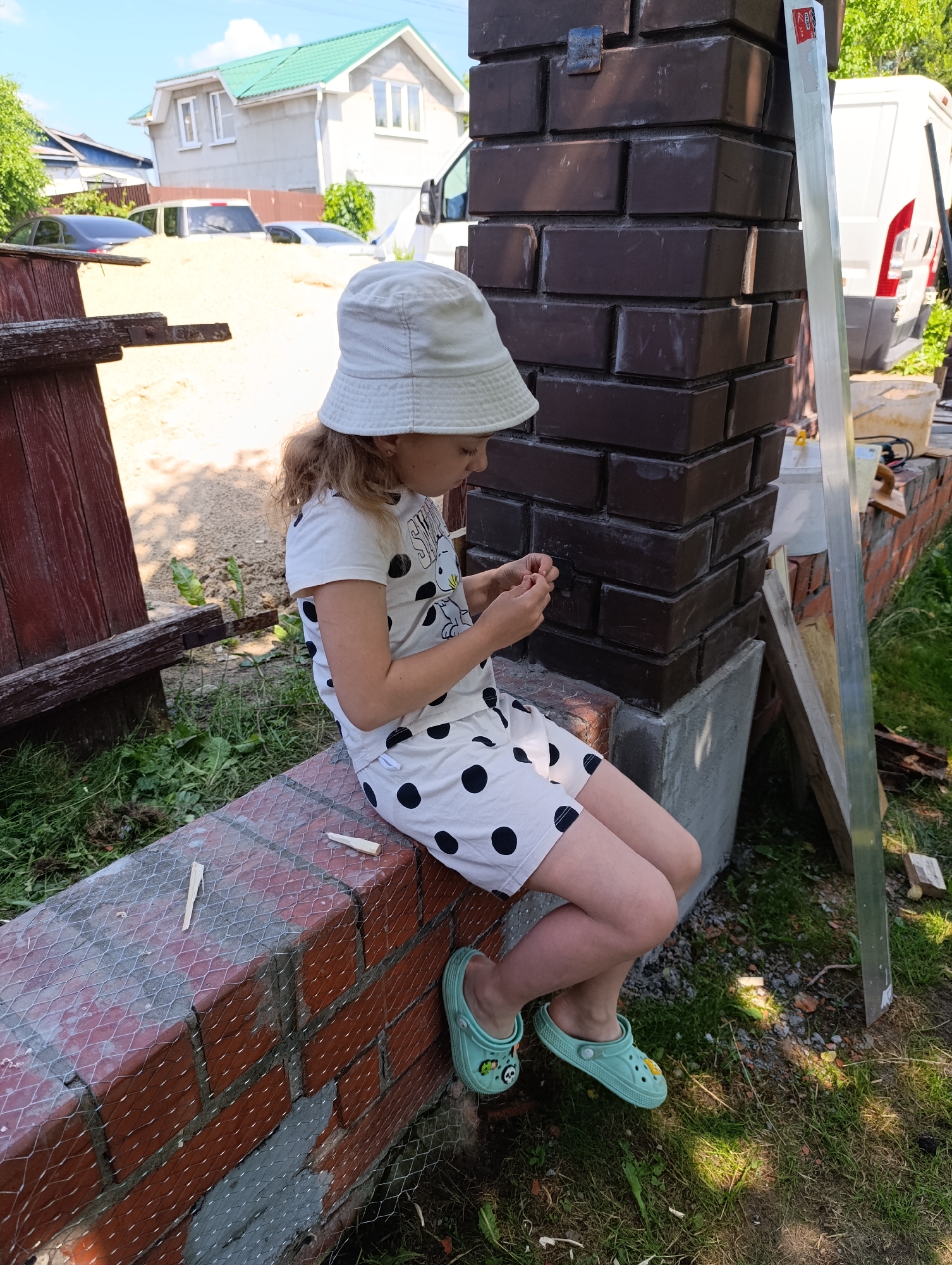 In an outdoor, daytime scene in Unknown, Unknown, a young girl wearing a white bucket hat and a white dress with black polka dots sits on a brick ledge. Her hands are engaged in a task, holding small items. The girl is wearing light green Crocs. The environment includes a brick pillar, grass, and a construction area with sand and building materials. In the background, a white van and a house under construction are visible.