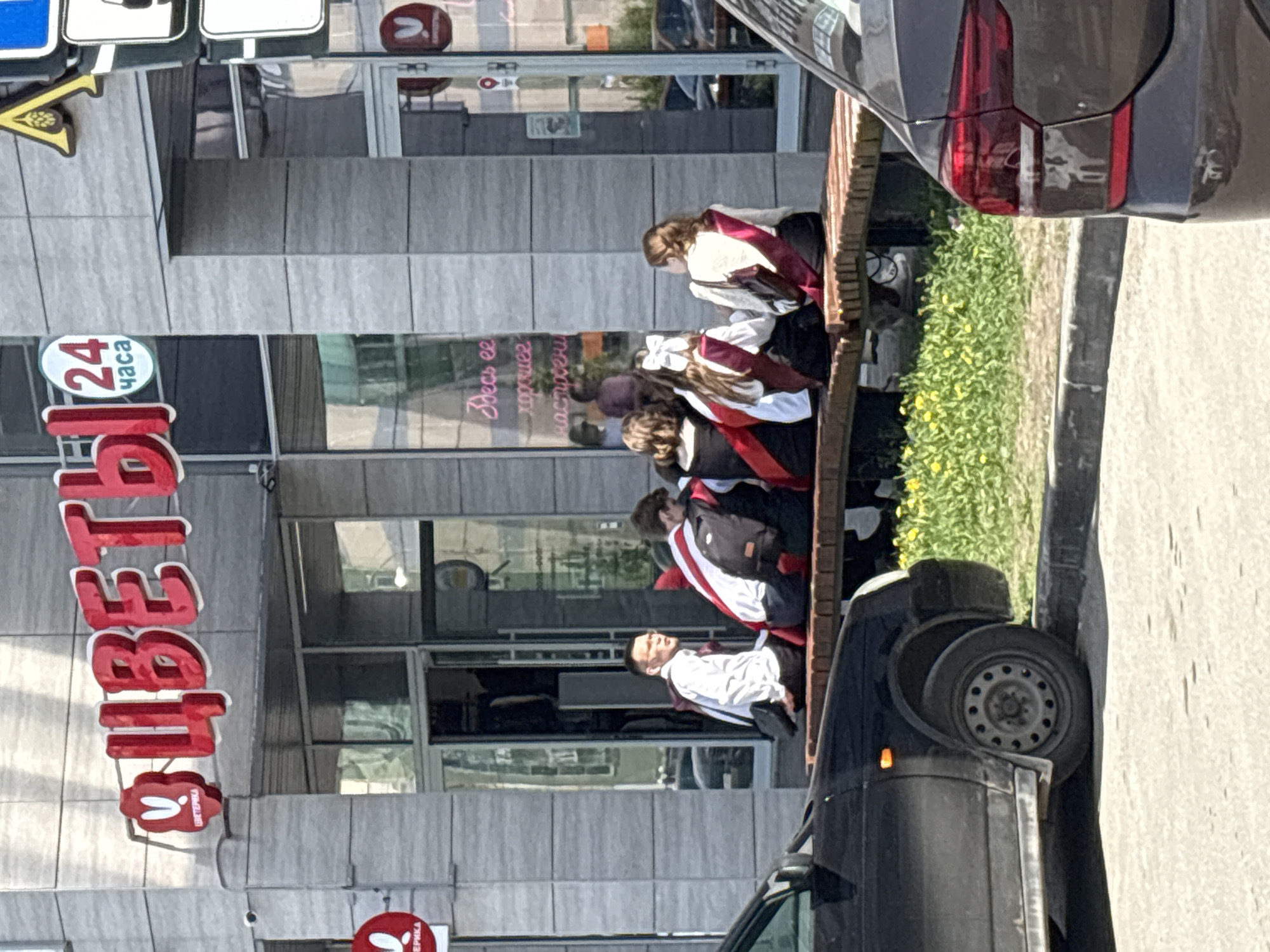The image shows a street scene in Perm, Russia, indicated by the Cyrillic lettering "ЦВЕТЫ" (flowers) on a store sign. Several young people, likely graduating students, are seated on a bench outside a building with reflective glass windows. They are wearing white shirts or blouses with red and black graduation sashes. The sign on the storefront window in Cyrillic indicates "Here it is...a good mood." Two cars are partially visible, one parked in front of the students and another on the right side of the frame. The bench faces a small strip of green vegetation. Street signs are visible in the upper left corner. A "24 часа" (24 hours) sign is next to the store sign.