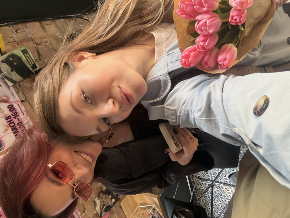 Two females are captured in a close-up selfie within an indoor setting. The individual on the left has reddish-brown hair, smiles at the camera, and wears red-tinted rectangular sunglasses, a dark jacket over a light top with a beaded necklace, and holds a light-colored smartphone. The individual on the right, with blonde hair, looks directly at the camera with a neutral expression. She wears a light blue trench coat over a white top and holds a bouquet of approximately ten pink tulips wrapped in brown paper. The background features an exposed brick wall adorned with various posters. One poster prominently displays "MARCH 8 / 13:00-16:00" and "PurrMačka." Another poster shows a black cat silhouette with text including "DORĆOL" and "PURRPICKLE," confirming the location as the Dorćol neighborhood in Belgrade, Serbia. A geometric tiled floor and a partial view of a table with a wooden display box are visible in the lower left background.
