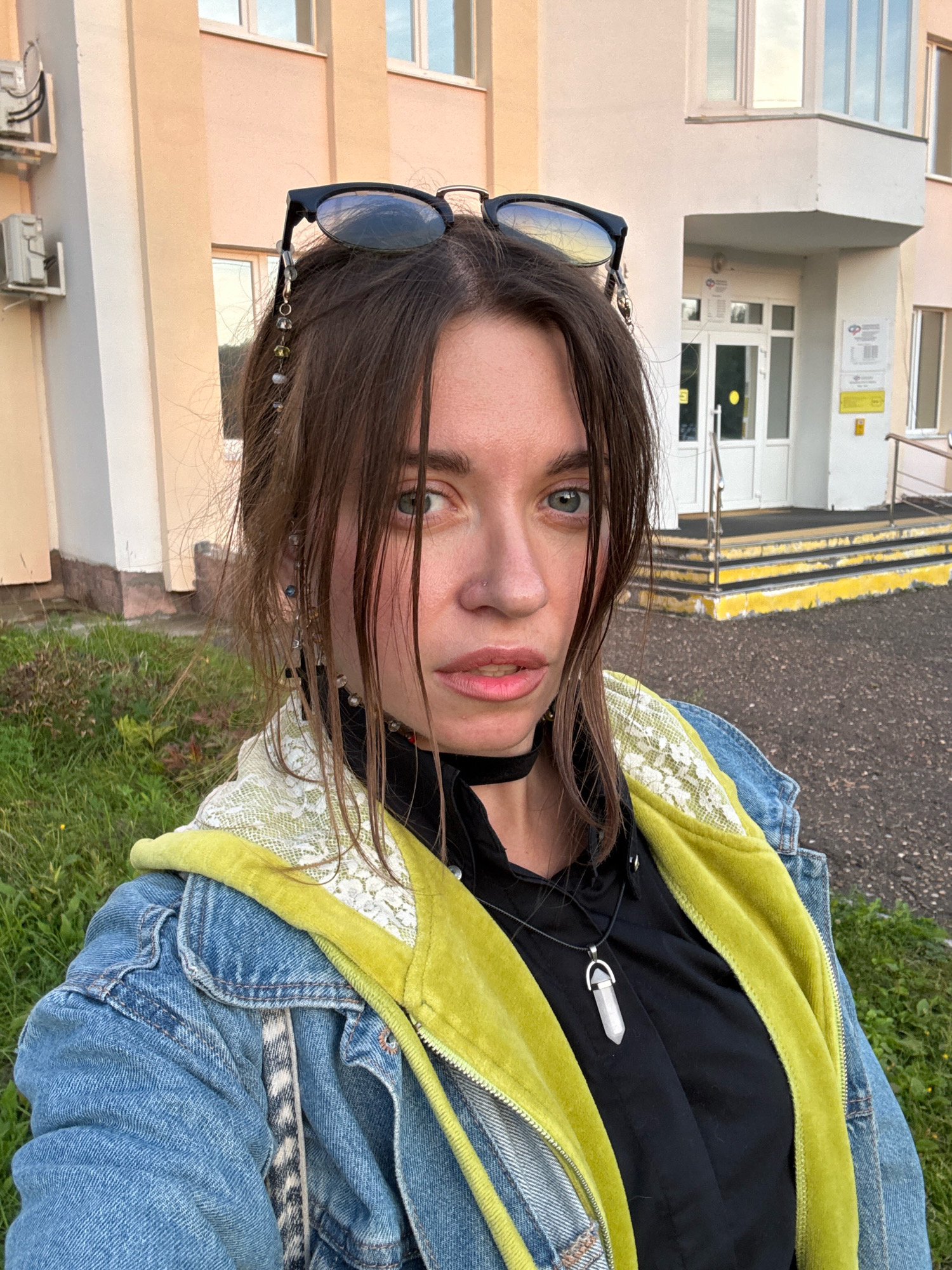 A medium shot depicts a young woman with brown hair and blue eyes posing for a selfie outdoors. She is wearing a denim jacket over a black shirt with a lime green hooded lining and a white lace trim. A black choker necklace with a silver pendant adorns her neck. Her sunglasses are perched on top of her head, with decorative bead strands attached to them, hanging down her hair.
The background shows a building with a light pink and white facade, featuring windows and an entrance with a yellow-accented staircase. Green grass and some gravelly ground are visible in the foreground. The overall lighting suggests it is daytime, possibly late afternoon or early evening, with the sun casting soft shadows. The environment appears to be an urban or suburban setting, possibly near an educational institution or administrative building.