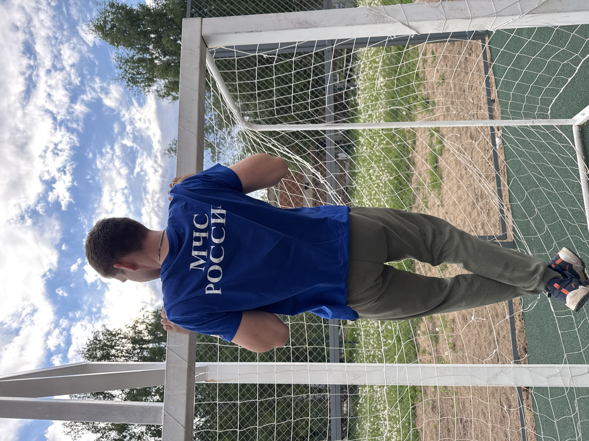 A man wearing a blue t-shirt with the Cyrillic inscription "МЧС РОССИИ" is positioned in front of a soccer goal in Вассята, Russia. The goal has a white net and a metal frame. The man is wearing green pants and athletic shoes. The background includes a cloudy sky and trees. The ground near the goal is visible through the net, showing grass and a textured green surface.