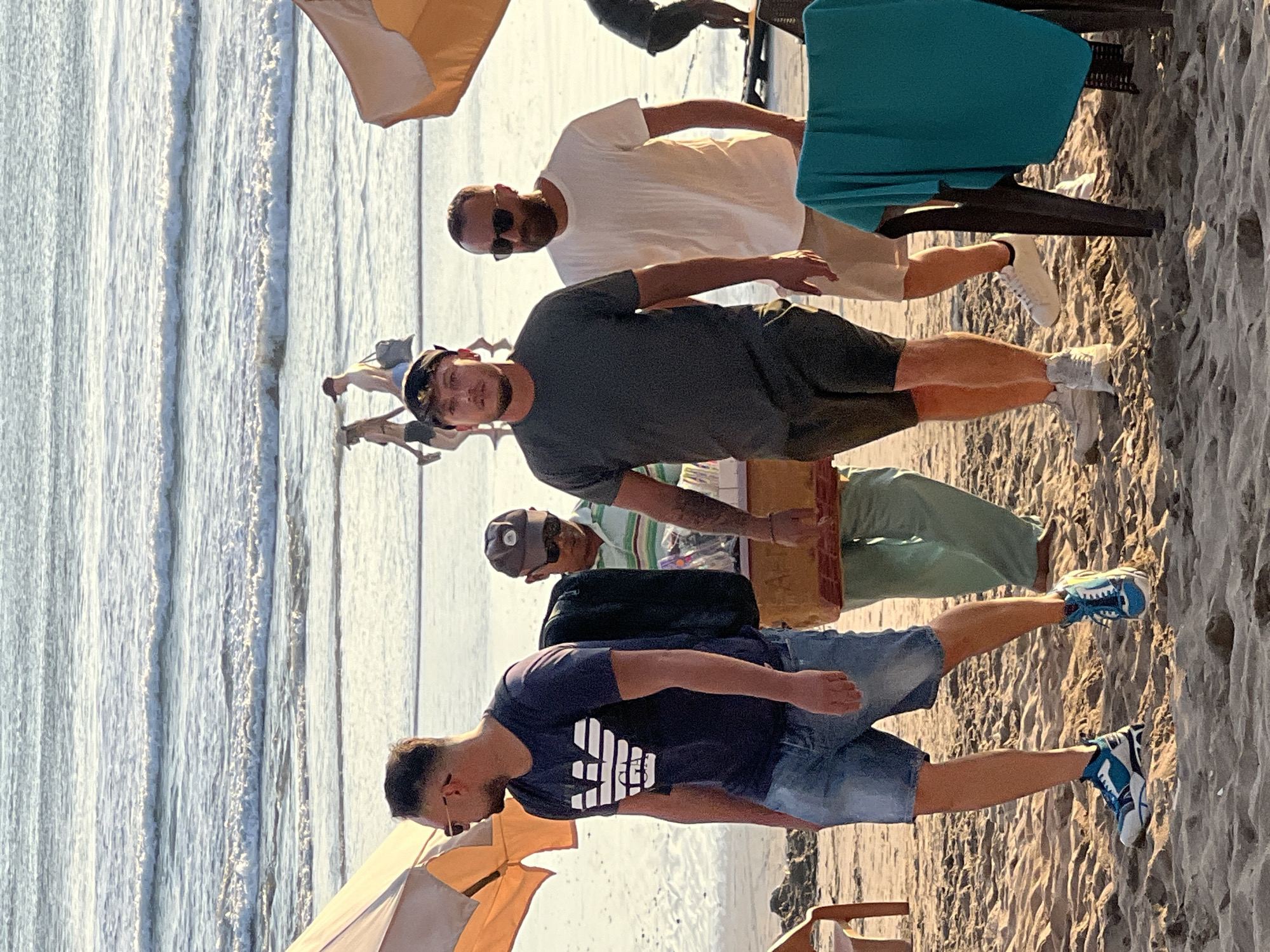 The image depicts a beach scene in Kuta, Indonesia, likely during the late afternoon given the low sun angle and warm lighting. In the foreground, four individuals are walking on the sand. The leftmost person is a man wearing a dark blue t-shirt with a white logo, denim shorts, and blue and white athletic shoes. To his right is another man in a dark grey t-shirt and dark shorts, wearing a black baseball cap. Behind them, a person in a green outfit and a dark baseball cap is carrying a wooden crate. To the right of the man in the grey t-shirt is a man in a white t-shirt and light-colored shorts, wearing sunglasses. In the background, the ocean with moderate waves is visible, and other people are walking along the shoreline. Two beach umbrellas are present, one on the left and one on the right, casting shadows on the sand. Beach chairs are also visible. The overall atmosphere suggests a casual and leisurely day at the beach.