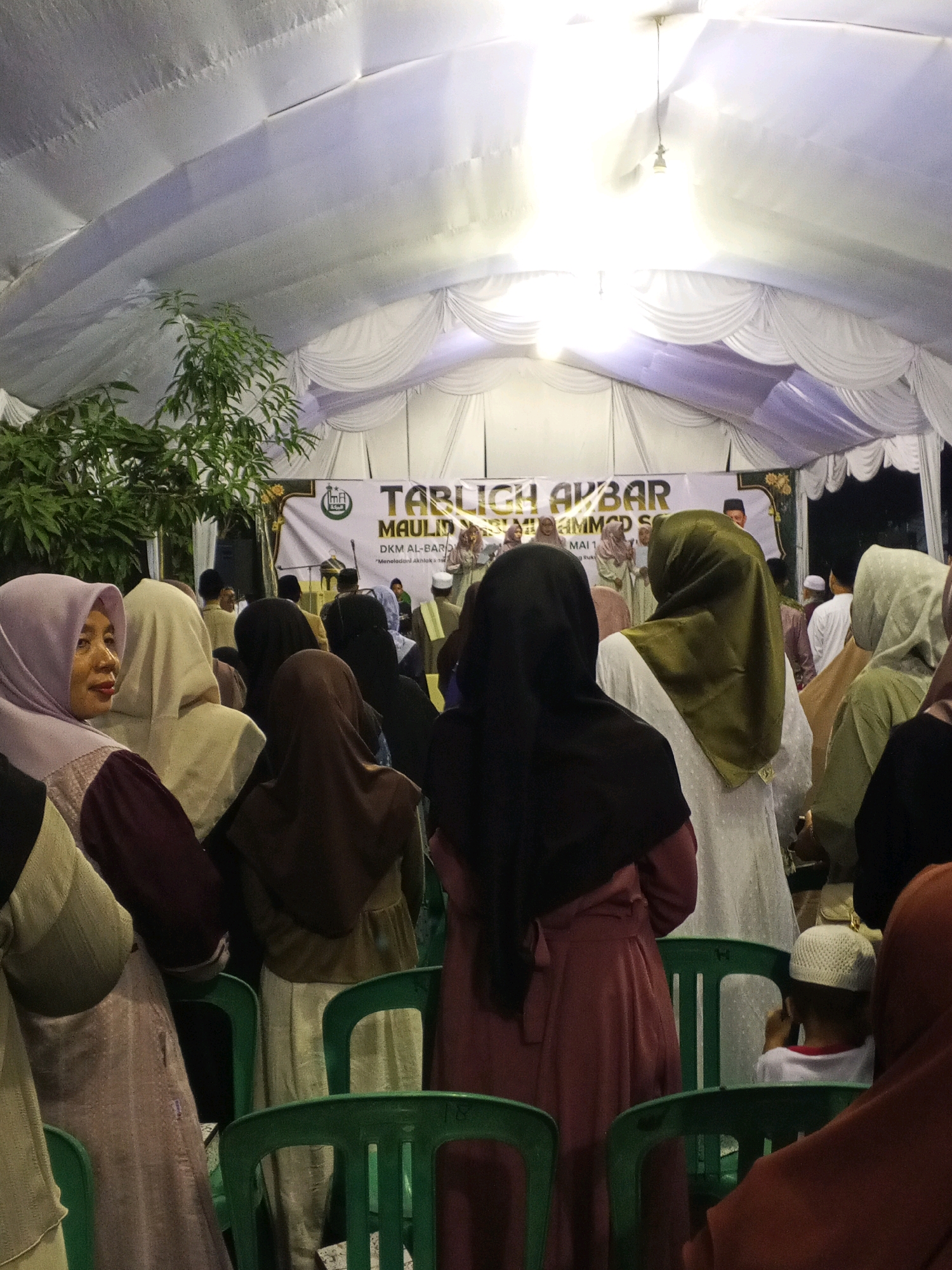 In Ciruas, Indonesia, a large tent structure serves as a venue for an outdoor event, possibly a religious gathering given the banner that reads "TABLIGH AKBAR MAULID NABI MUHAMMAD SAW" and "DKM AL-BAROKAH". Inside the tent, a crowd of people, predominantly women wearing hijabs of various colors including purple, beige, brown, and green, are seated on green plastic chairs. Some individuals are standing closer to a raised platform or stage area, where a banner and a microphone are visible. The environment appears to be set up for a formal event, with white fabric draped to form the tent's walls and ceiling, illuminated by overhead lighting. The overall scene depicts a community gathering under a temporary structure.