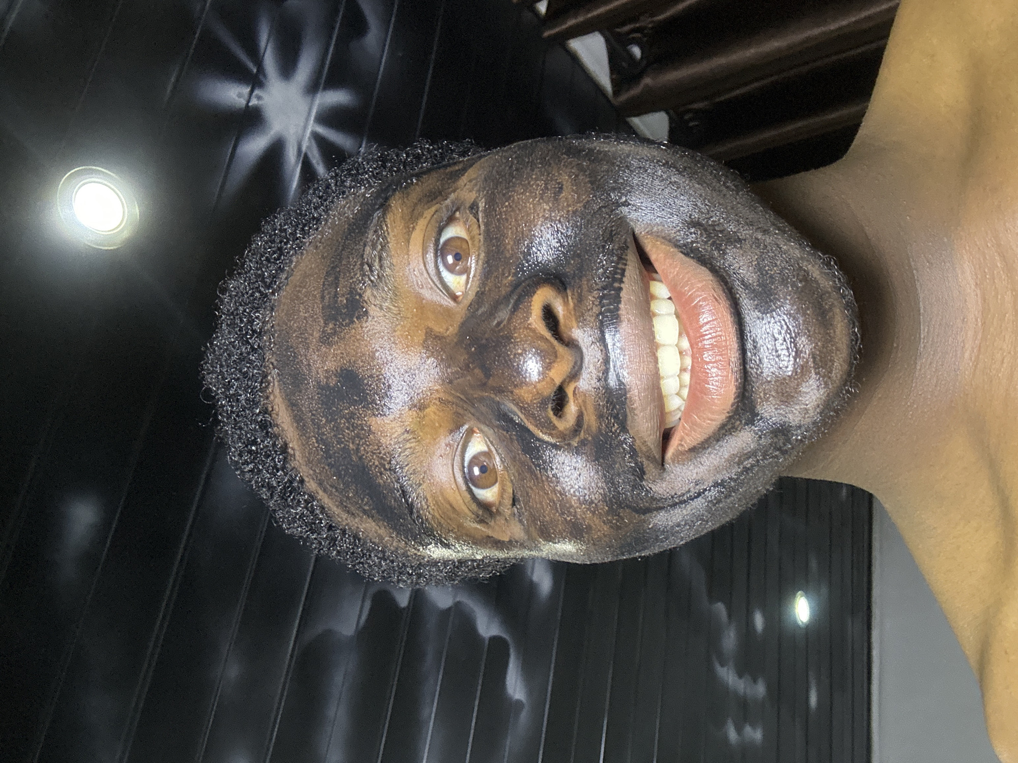 A close-up, upward-angled shot depicts a smiling person with a dark, textured substance applied to their face, possibly a facial mask. Their hair is short and curly. The person has brown eyes and their teeth are visible. The background consists of what appears to be a dark, possibly paneled, wall or surface with light reflections. There is no information in the image that confirms the location in the city of Bukuru, Nigeria.