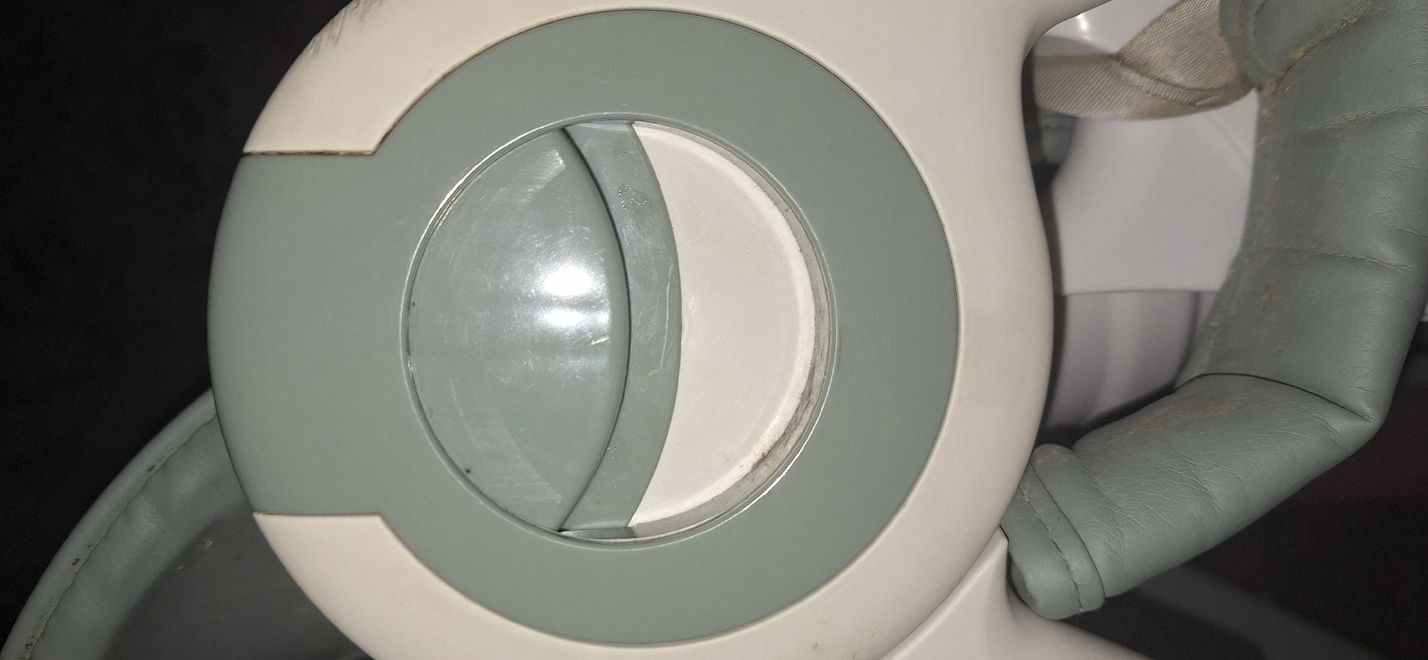 The image is a close-up, low-angle shot of a light-colored plastic or metal device with a circular opening. The device has a light gray outer rim and a white inner circular area. Within the white circle, there is a slightly darker gray, curved plastic piece, suggesting a movable part or a handle. The surface of the gray plastic shows several light scratches and scuff marks, indicating wear and tear. There are also some dark specks, possibly dirt or debris, visible on the gray and white surfaces, particularly around the edge of the inner gray piece. The lower portion of the image reveals a textured, light gray material, likely padded leather or a similar synthetic, with visible stitching and some dirt. A beige strap is also partially visible. The background is dark and out of focus, with some brighter reflections on the device's surface. This object appears to be part of a larger piece of equipment, possibly a baby bouncer or a similar child-related item. The image does not contain any elements that would indicate its location within the city of Bila Tserkva, Ukraine.
