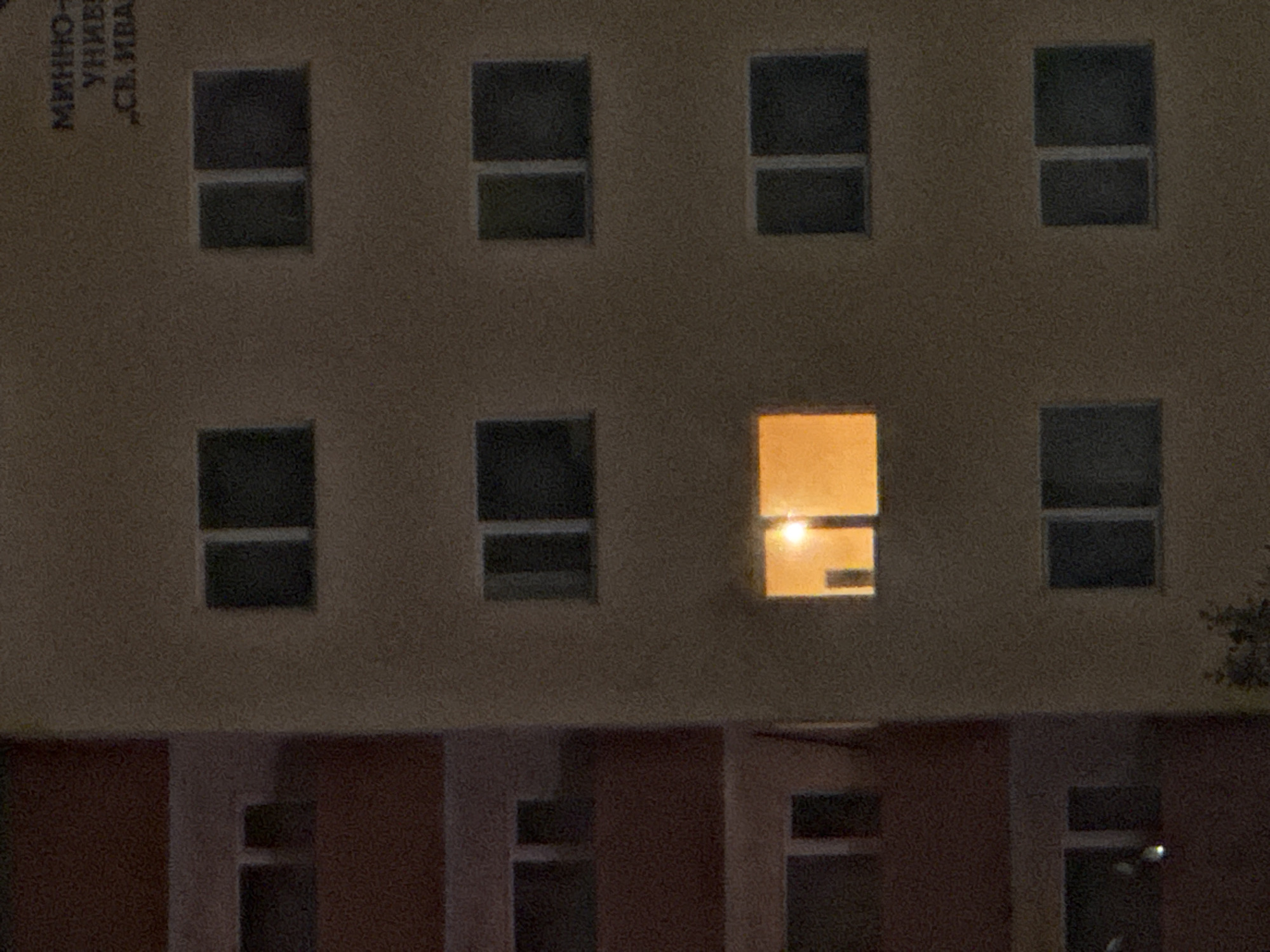 This nighttime photograph shows a building in Sofia, Bulgaria. The building is multi-storied and has windows arranged in a grid pattern. Most of the windows are dark, except for one which is illuminated with a warm, golden light. At the top of the building is a name written in Bulgarian, identifying the building as belonging to Sofia University "St. Kliment Ohridski". There are other buildings in the foreground. A tree branch is visible on the right side of the image.