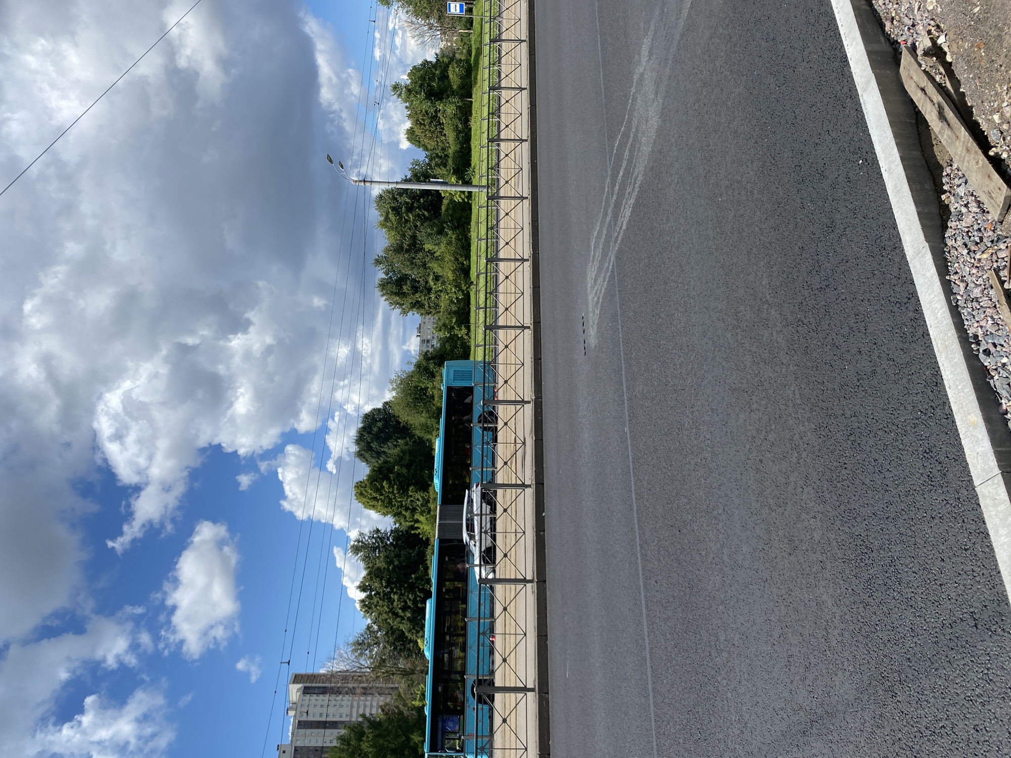 An outdoor, eye-level, medium shot captures a bright, partly cloudy day in an urban environment, likely Saint Petersburg, Russia, given the architectural style and prevalent bus design. In the foreground, a freshly paved asphalt road dominates the lower half of the image, with a concrete curb and gravel lining the edge.
In the midground, a long, articulated, bright turquoise bus is parked parallel to a metal fence. The bus windows are tinted, and some passengers are visible inside. To the left of the bus, a white car is parked. The fence separates the road from a grassy area with trees and bushes. In the background, a multi-story residential building with a light-colored facade stands behind the trees. A street lamp and overhead power lines are visible against the sky. The sky itself is a vibrant blue, interspersed with large, fluffy white clouds. The overall impression is one of a typical city scene on a pleasant summer day.
