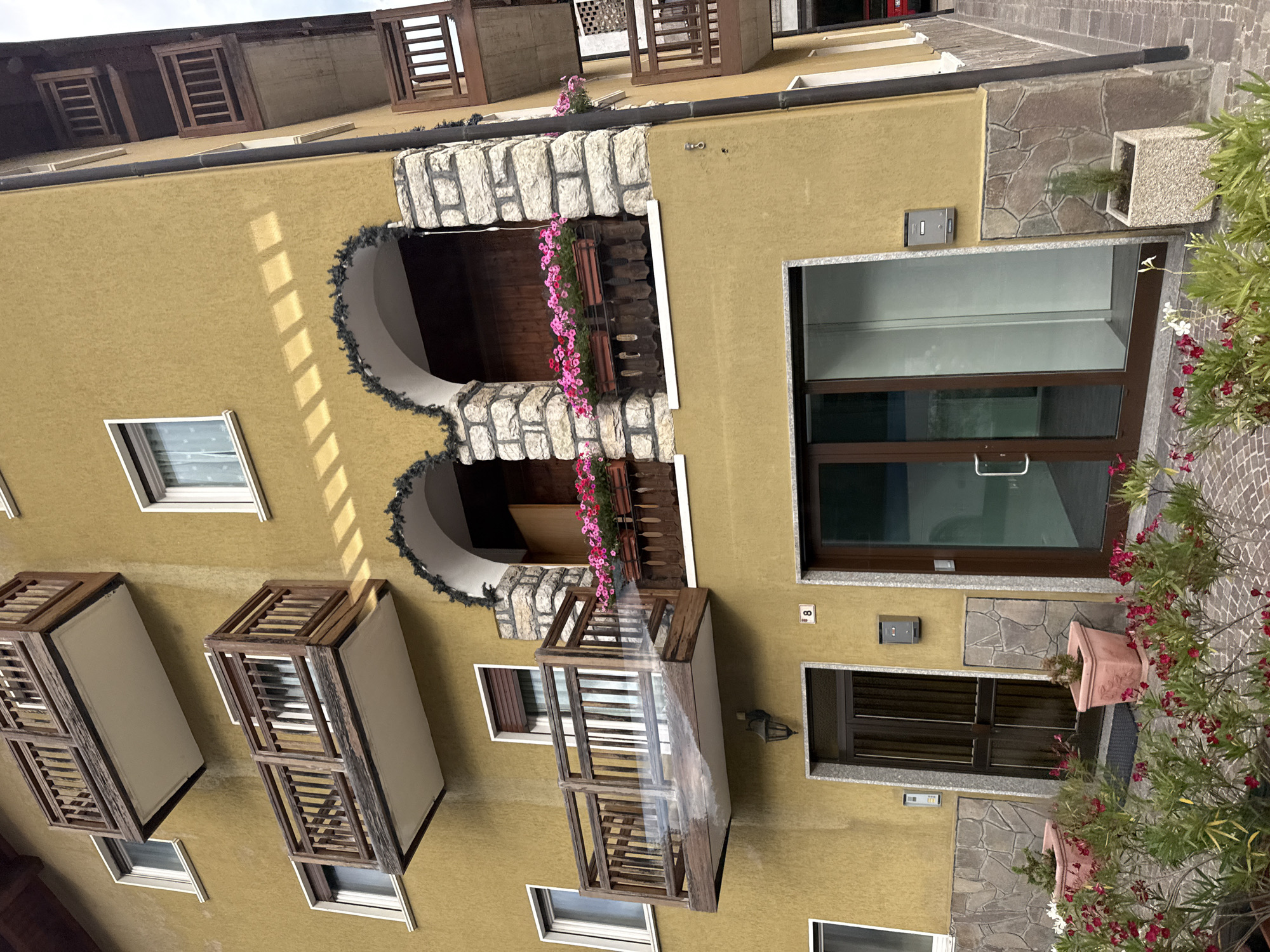 The image shows the exterior of a multi-story building in Cavedago, Italy, with a mustard-yellow stucco facade. The building features several wooden balconies with railings, some of which are adorned with planters filled with pink flowers. Two arched openings with stone accents are visible on the second floor, also decorated with planters. A dark brown door is situated on the ground floor, next to a glass-paneled entrance. Potted plants and flowering shrubs are arranged in front of the building. The scene is captured from a slightly upward angle, with the building's corner prominently displayed.